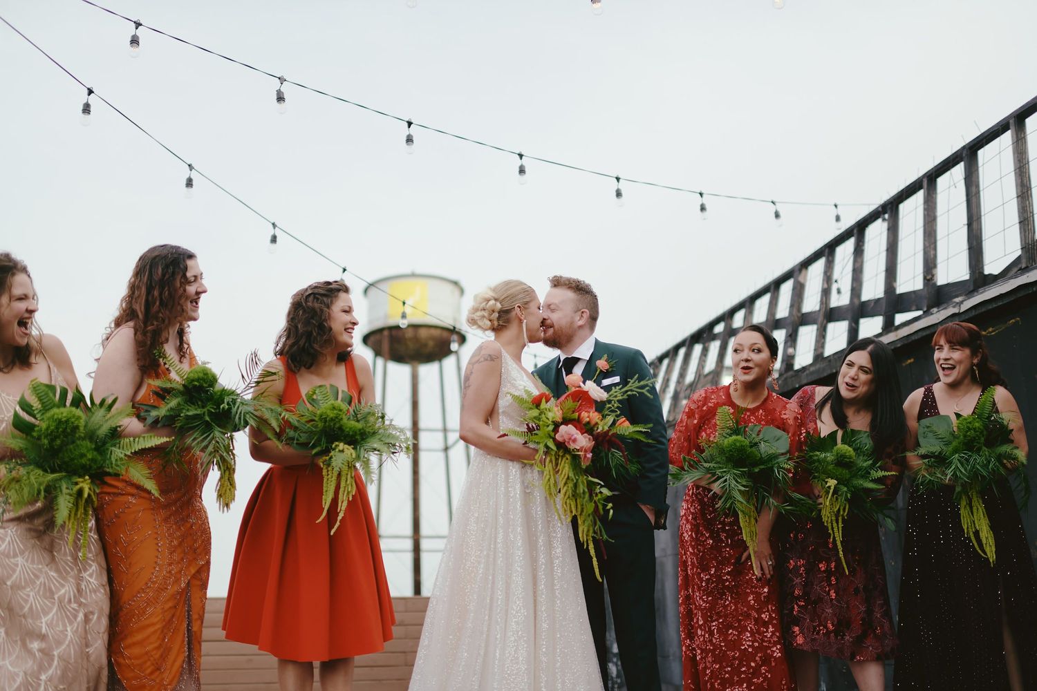 A wedding celebration on a rooftop deck with bridesmaids in colorful dresses holding tropical greenery bouquets.
