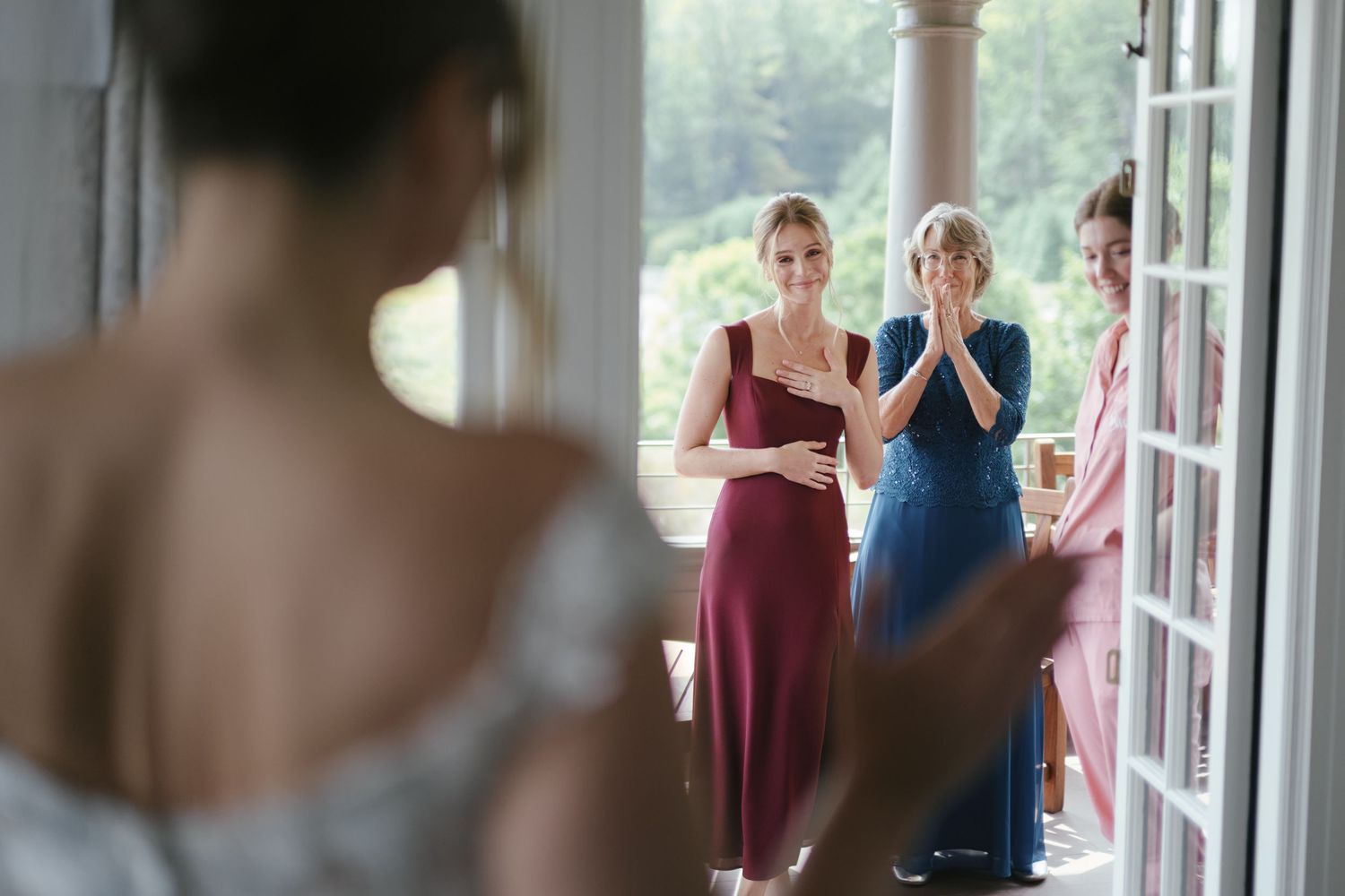 Bridesmaids in burgundy and blue dresses react emotionally as bride reveals her wedding gown during bridal preparation.
