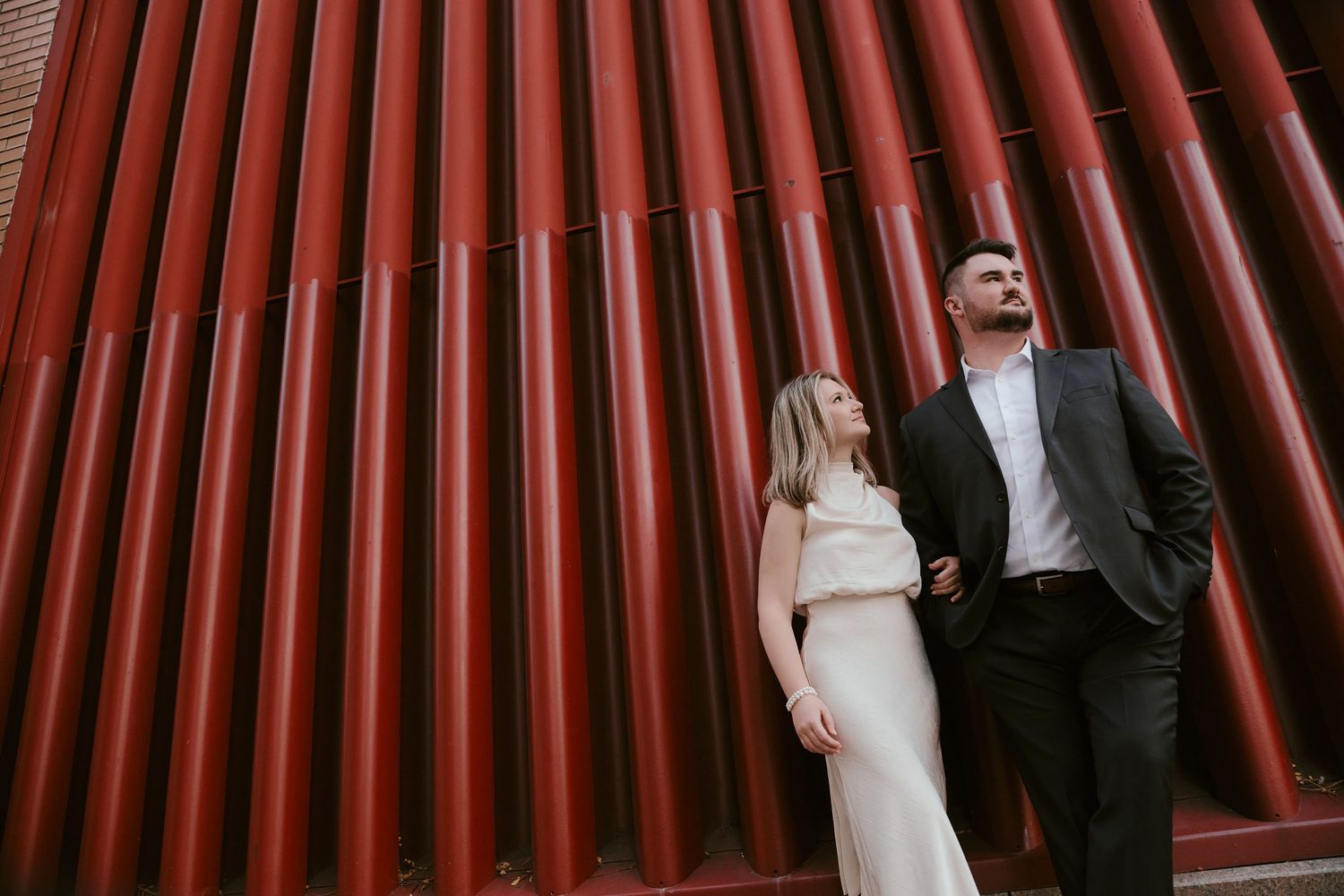 Couple poses in front of red cylindrical pipes, woman in white dress, man in dark suit, looking upward.