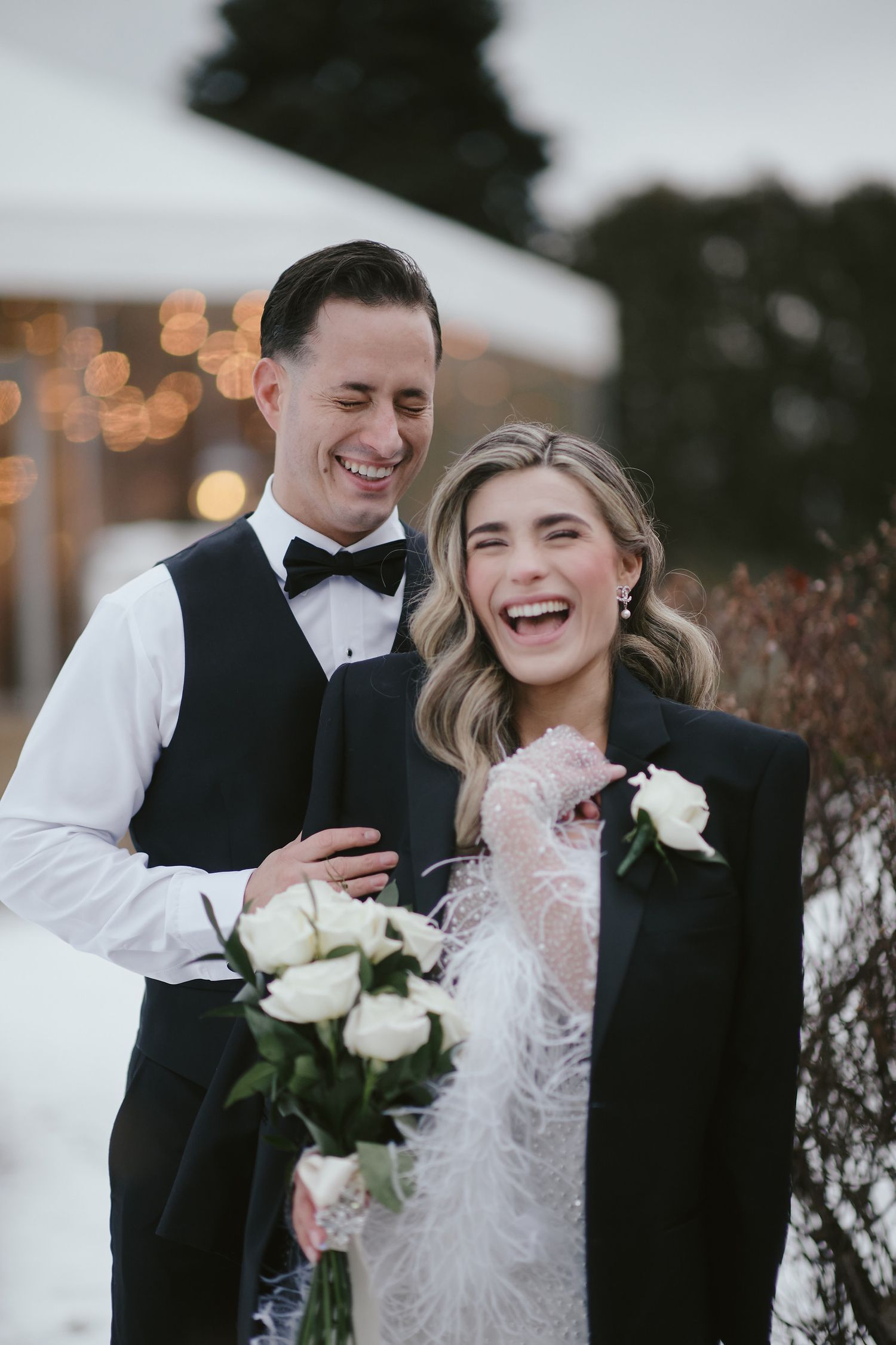 A joyful wedding couple sharing a candid moment outdoors with white roses and feather accents.