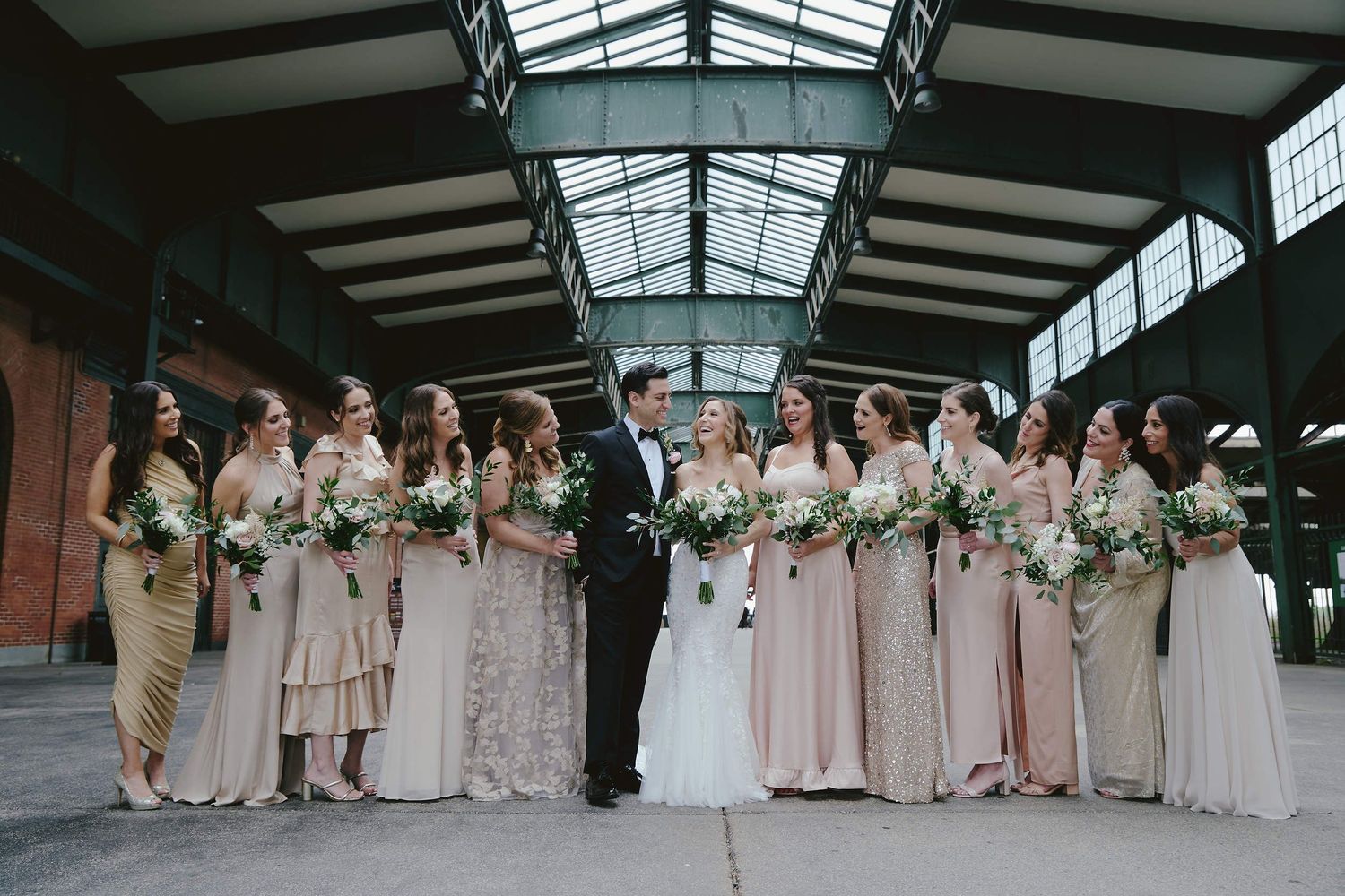 Wedding party poses together under covered walkway in neutral tones with white floral bouquets.