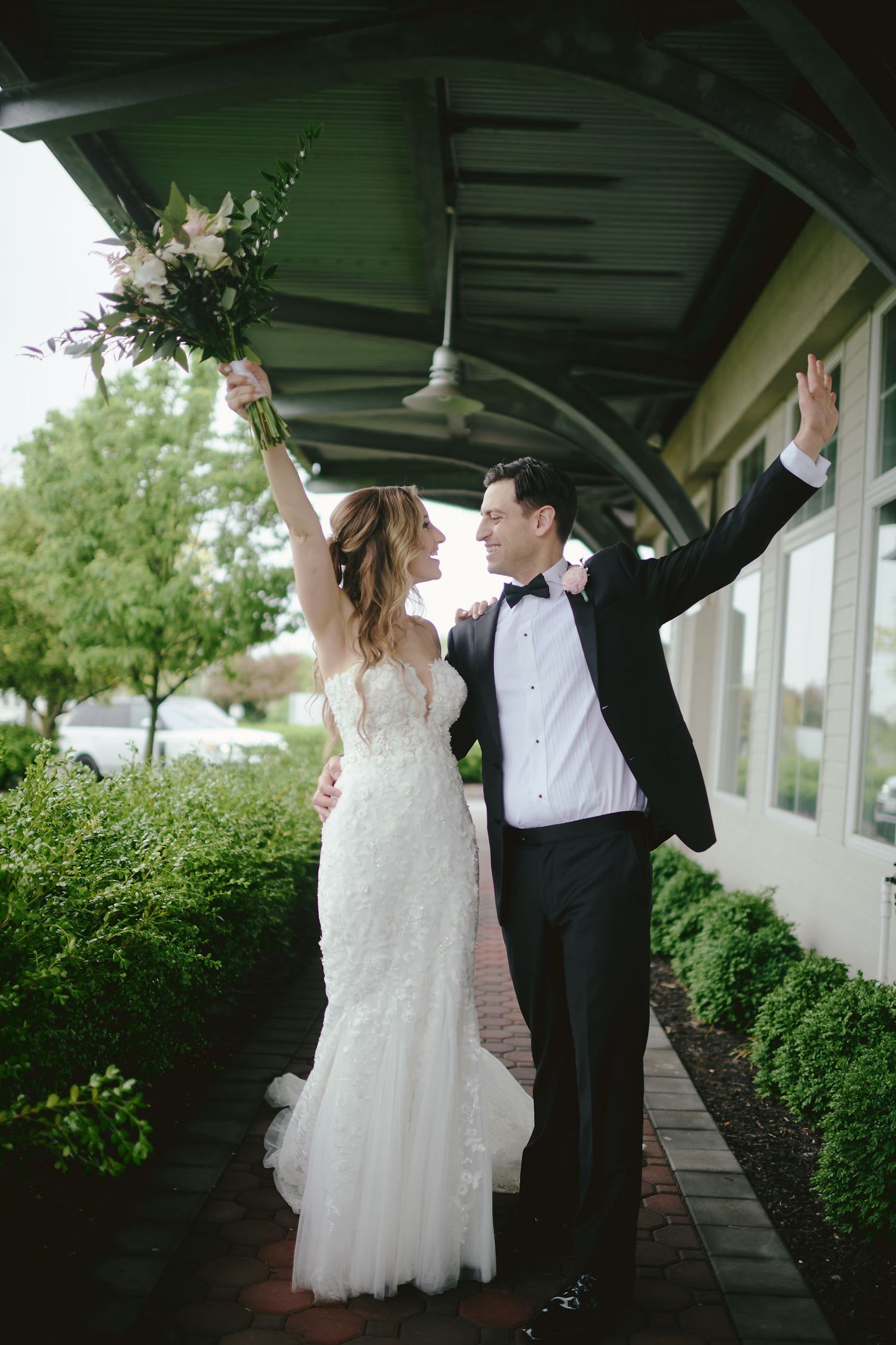 A joyful couple in formal wedding attire celebrate under a covered walkway with raised arms and a floral bouquet.