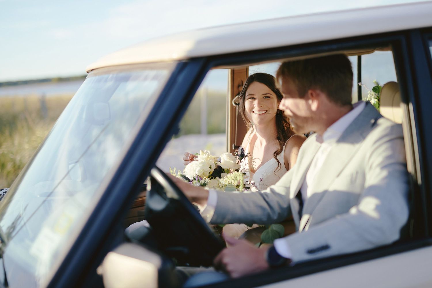 A couple sits in a vintage car with a bouquet of flowers while enjoying a sunny day together.