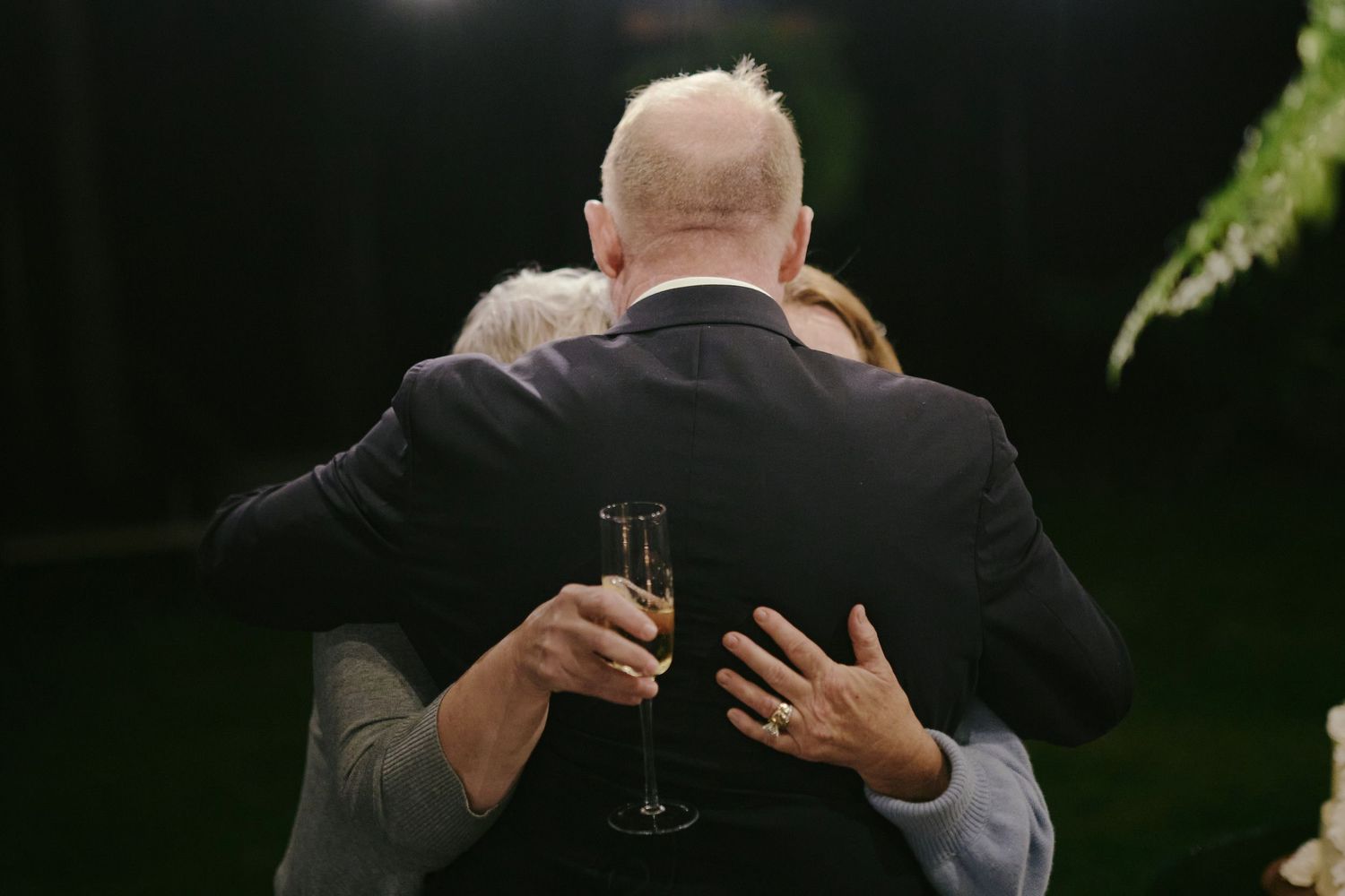Two people embrace while one holds a champagne glass during what appears to be an outdoor evening event.