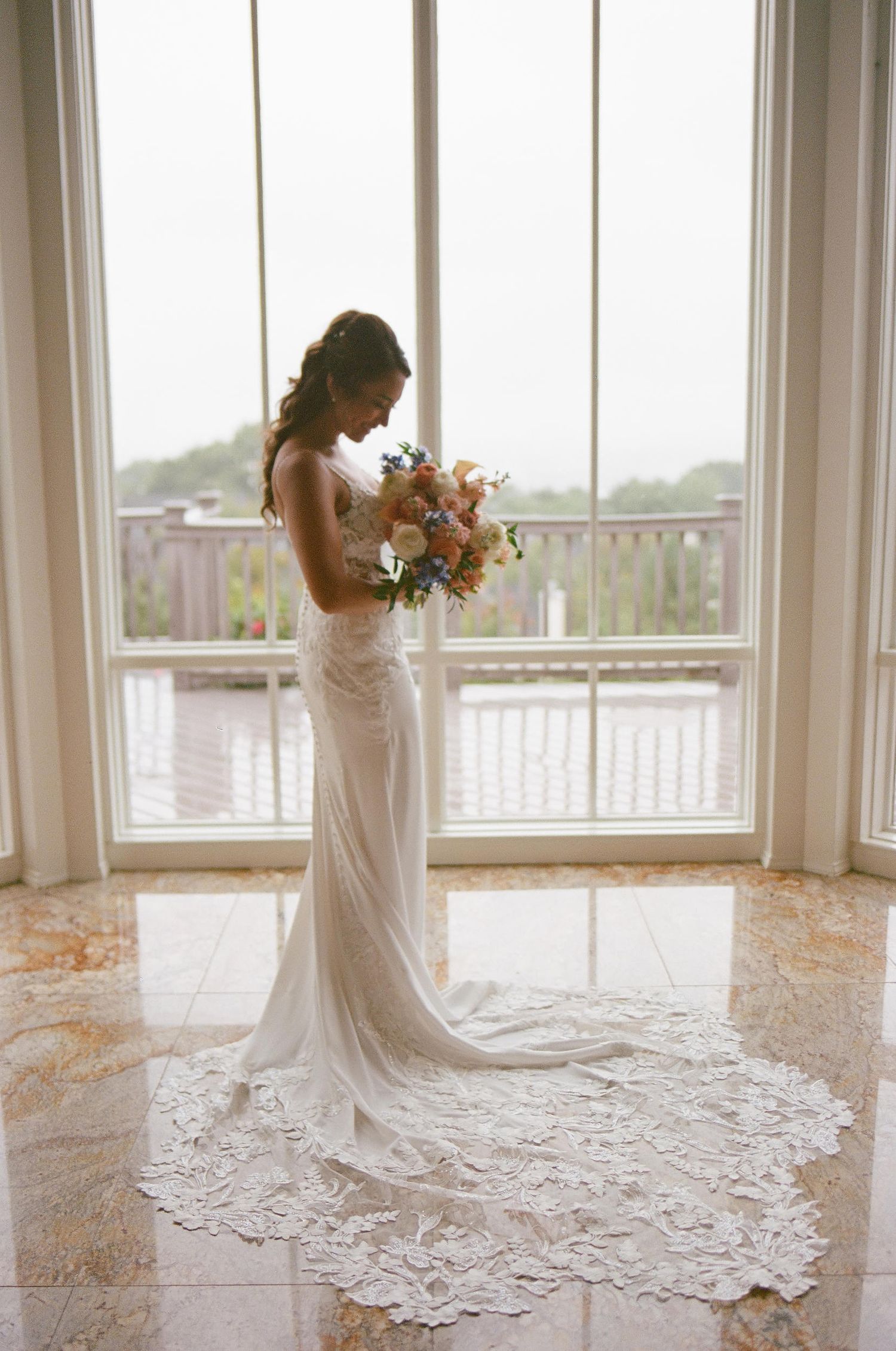 Elegant bride in lace train wedding gown silhouetted against bright window while holding floral bouquet.