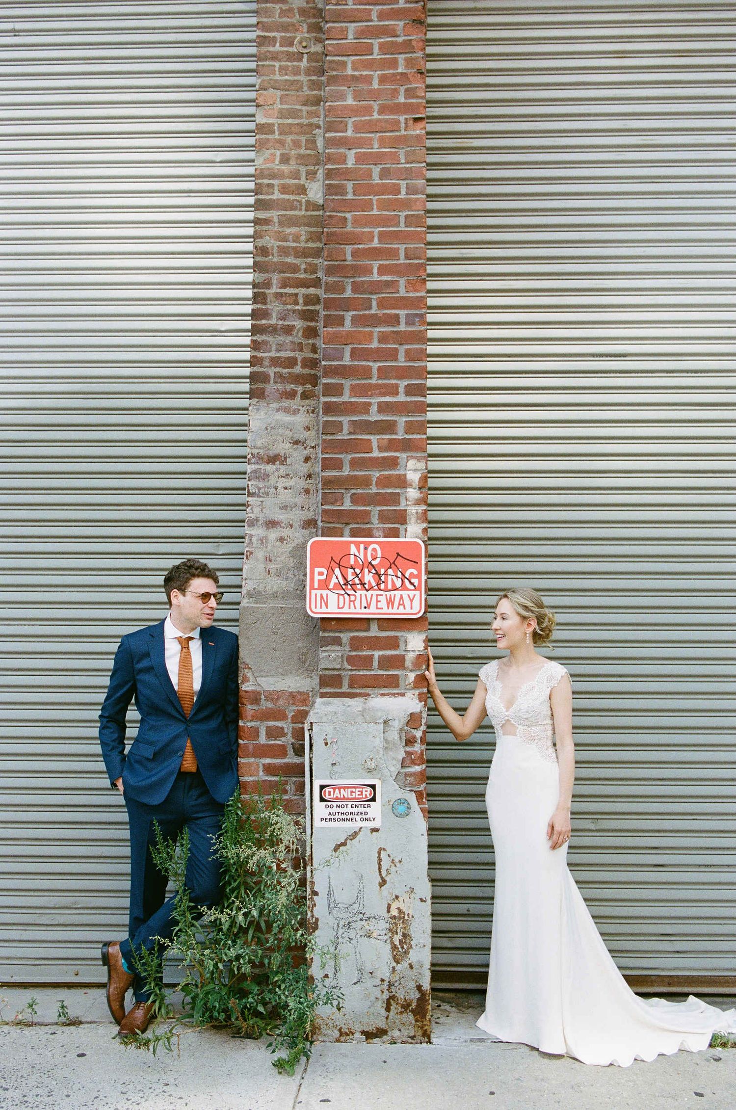 Wedding couple poses playfully against industrial brick wall with no parking sign.