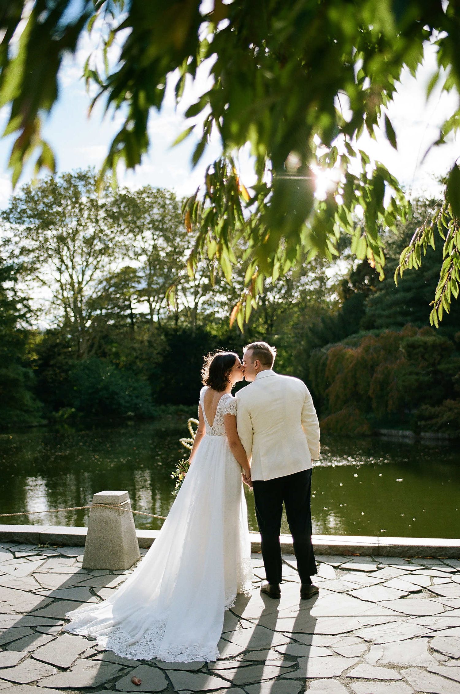 A couple shares a romantic moment on a stone terrace overlooking a tranquil pond, framed by willow leaves at sunset.