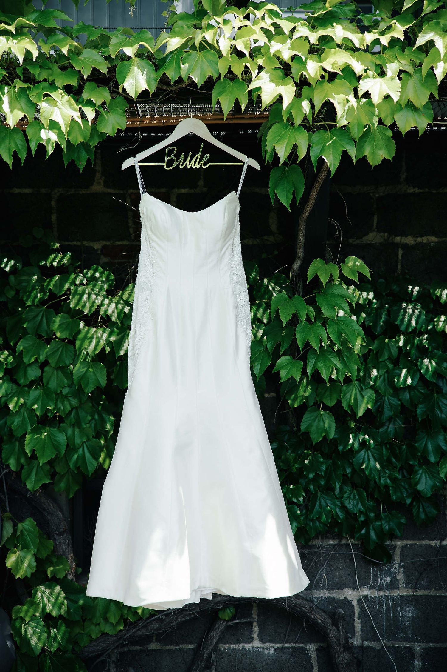 A white wedding dress hangs on a personalized bridal hanger against a backdrop of lush green ivy leaves on a wall.