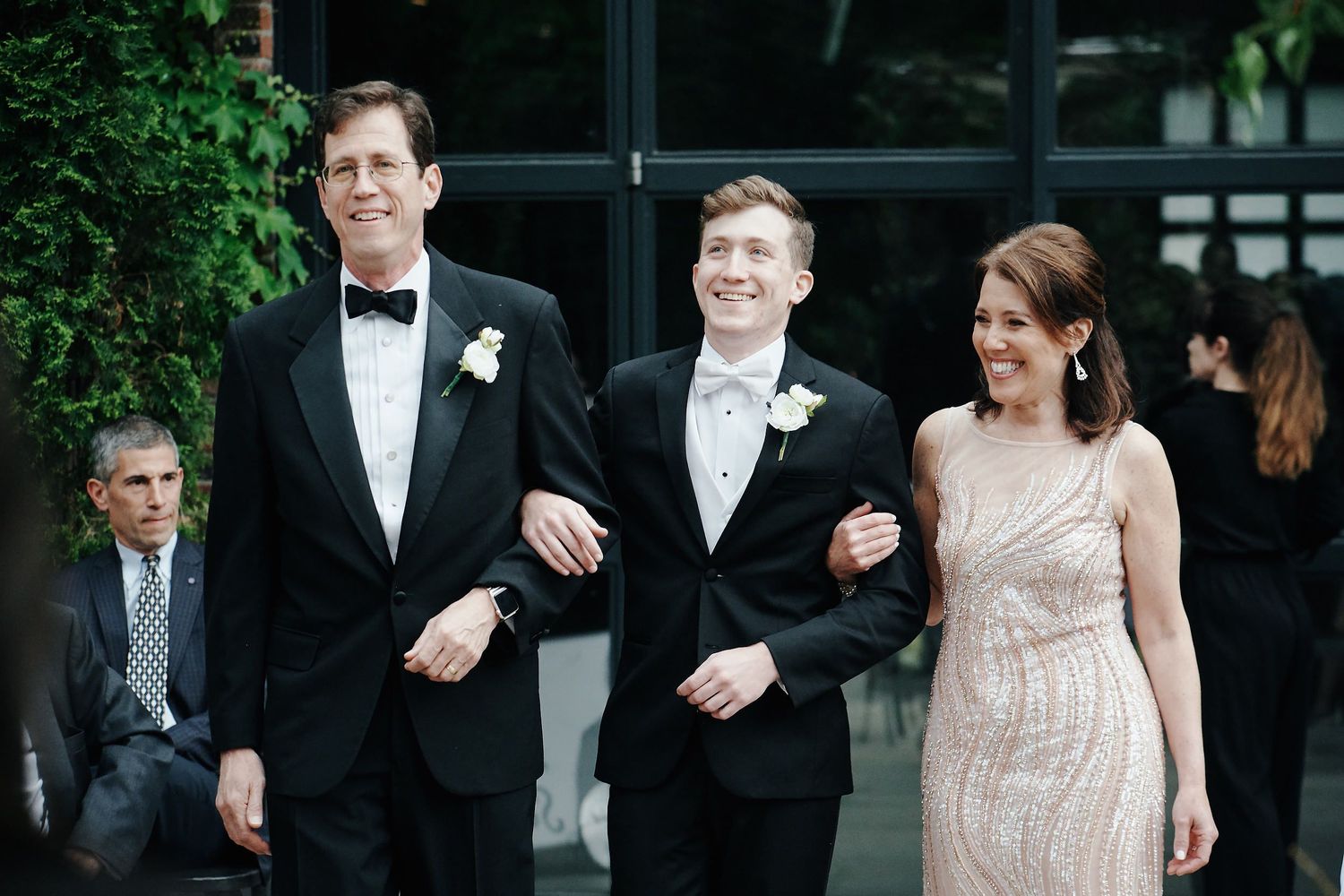Wedding party members dressed in black tuxedos and a soft pink gown walk arm-in-arm during an outdoor ceremony.