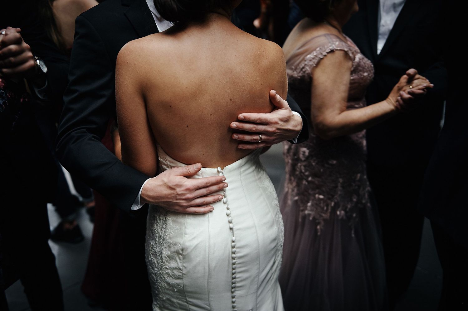 A close-up view of a wedding dress with buttoned detailing down the back as guests dance at a reception.