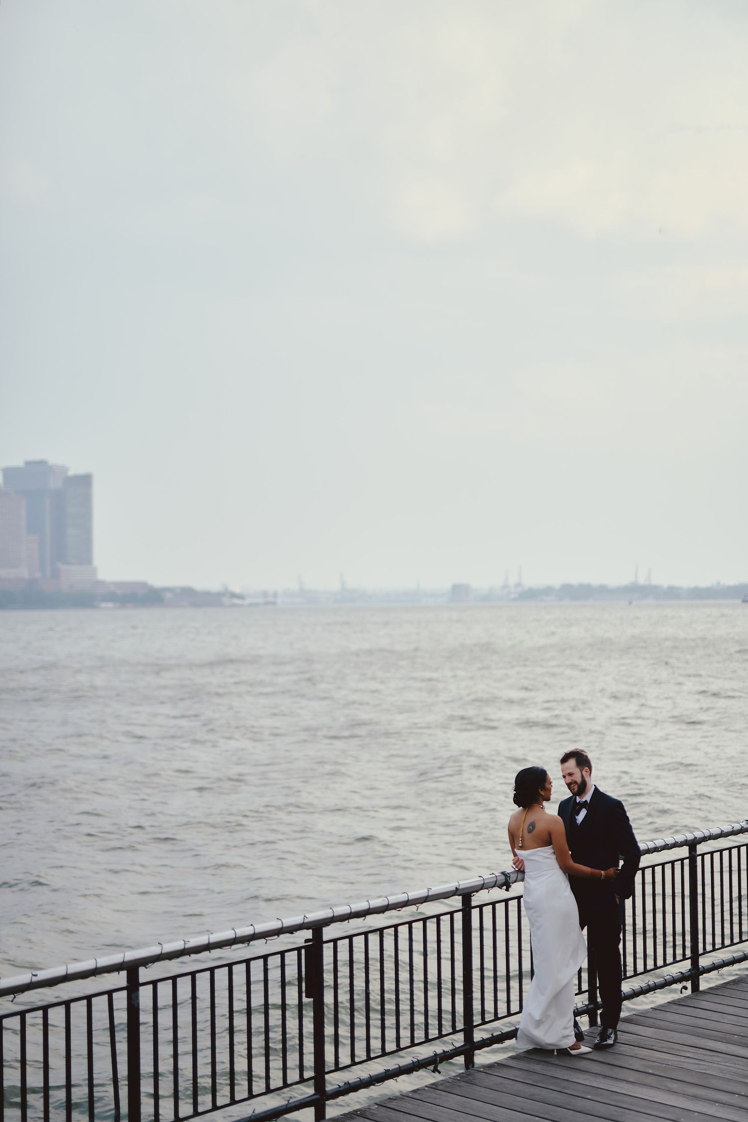 A couple shares an intimate moment on a wooden boardwalk overlooking a misty river with city buildings in the background.