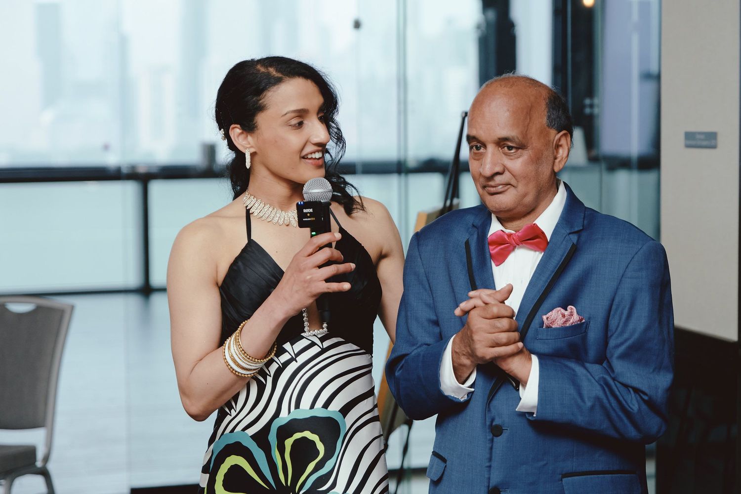 Two people at a formal event share a lighthearted moment while speaking into microphones in an indoor venue.