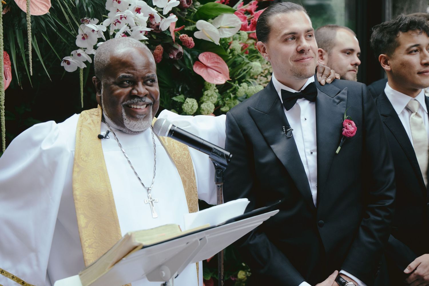 A clergy member in white robes and gold stole smiles while holding paperwork during a formal ceremony with attendees.