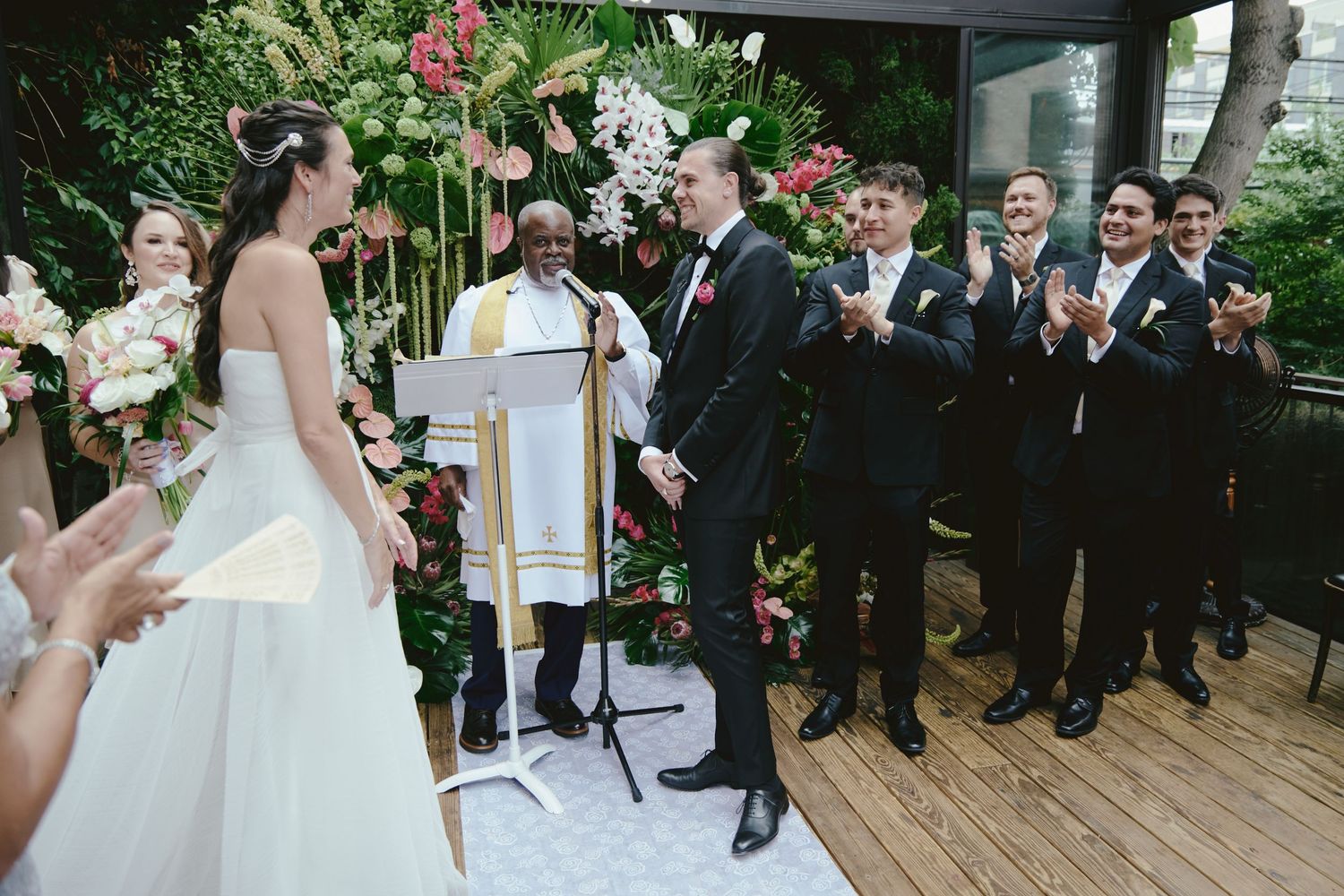 A wedding ceremony takes place on a wooden deck with floral arrangements and guests gathered around.