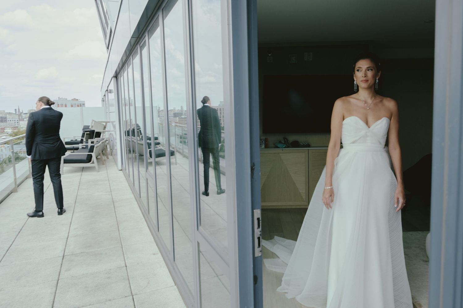 A person in a flowing white gown stands in a high-rise venue while suited figures observe the city from a balcony.