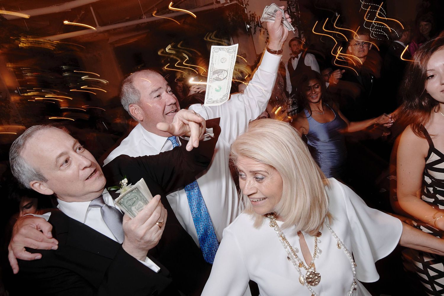 Partygoers in formal attire celebrate with money and enthusiasm on a dance floor at a nighttime event.