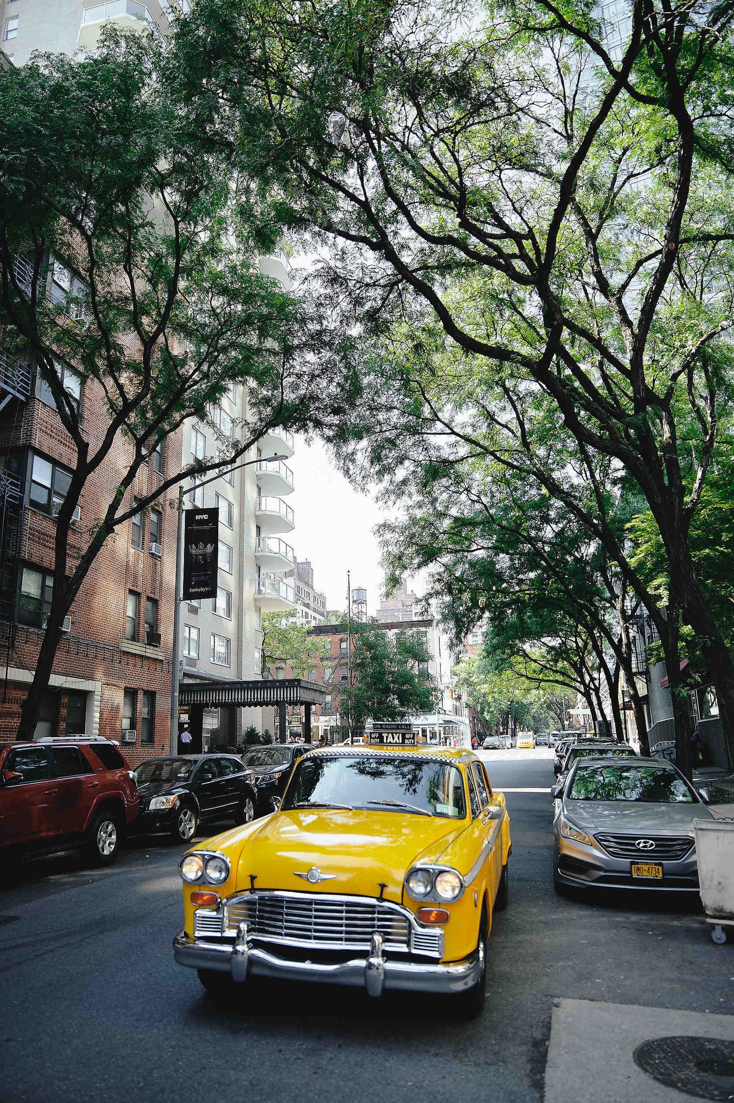 A bright yellow vintage taxi cab parked on a tree-lined New York City street with brick buildings in the background.
