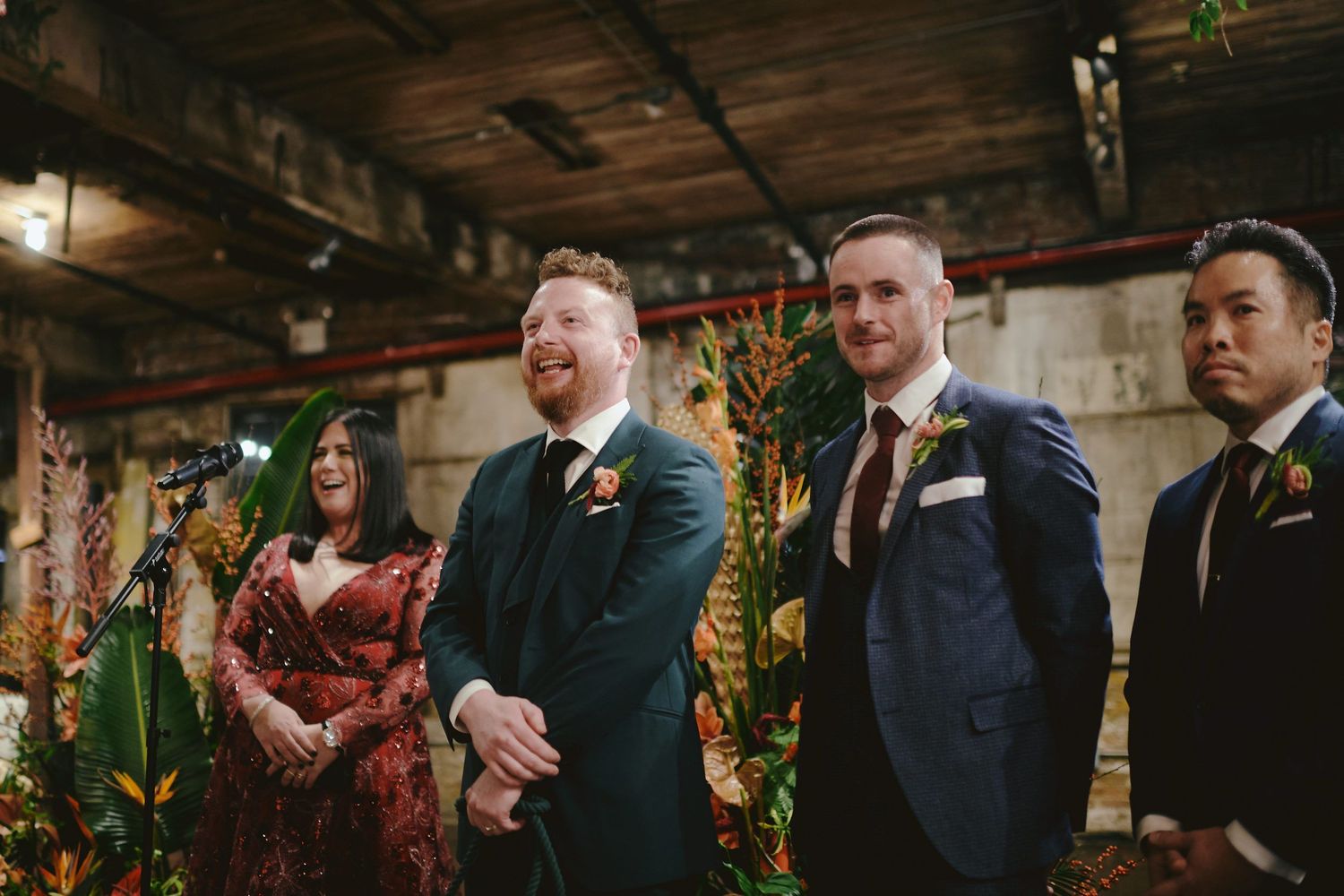 A wedding ceremony taking place in a rustic indoor venue with exposed wooden beams and decorative greenery.