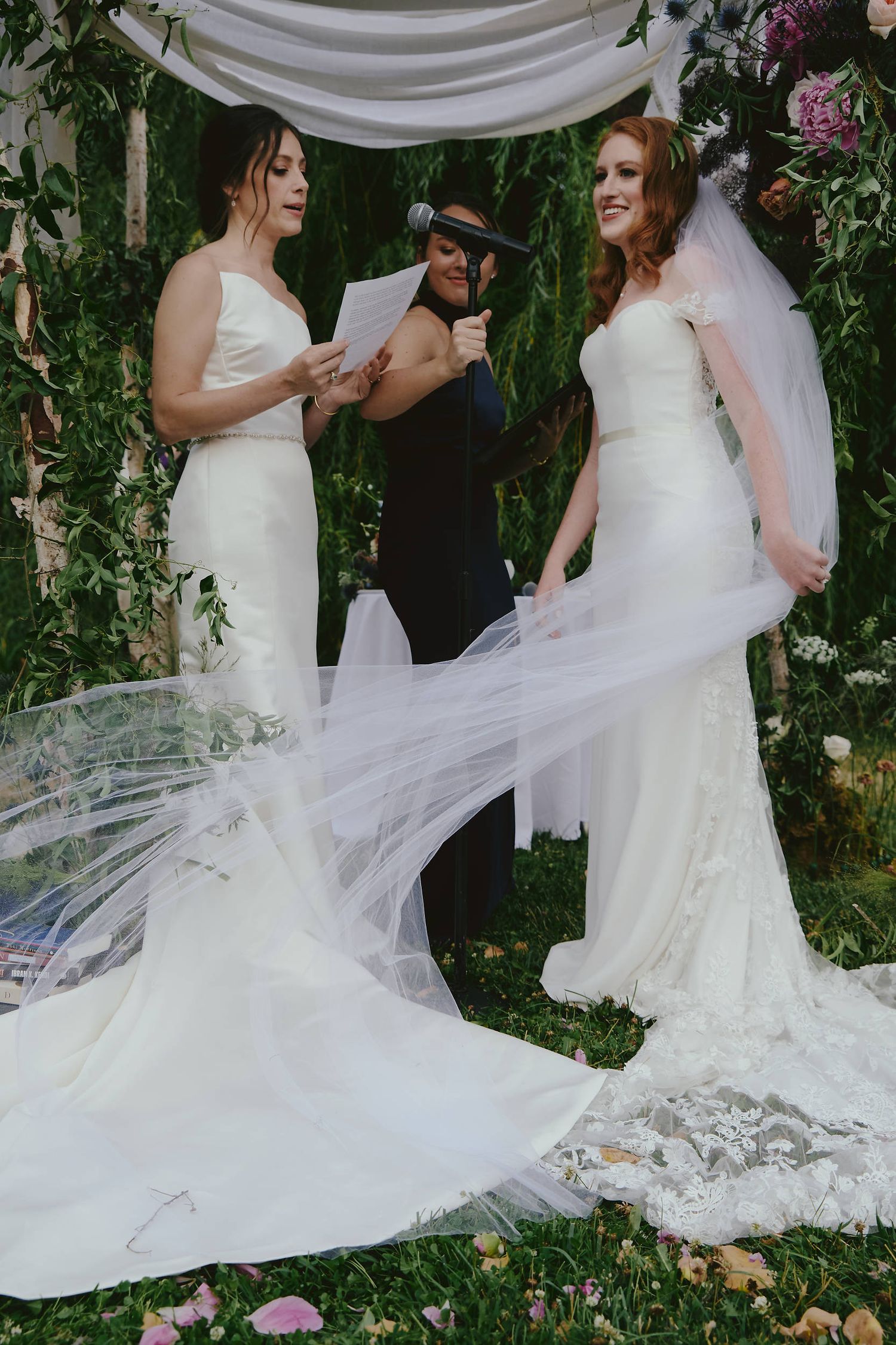 Two people in elegant white wedding gowns stand together under a floral archway during an outdoor ceremony.