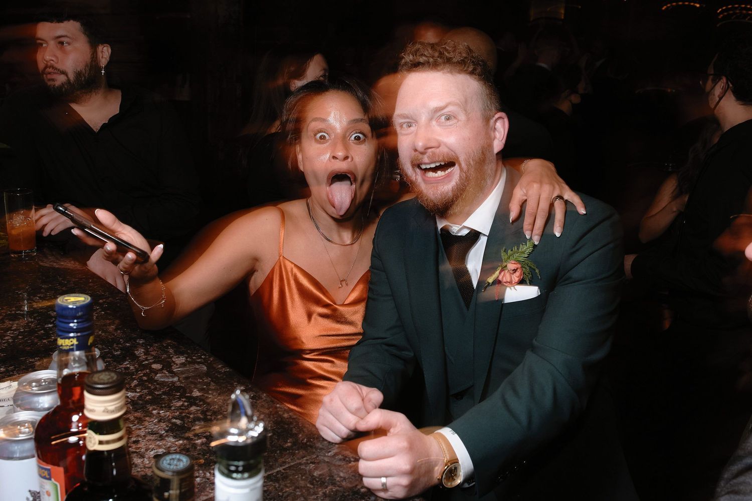 Two animated partygoers share an enthusiastic moment together at a bar, with drinks visible on the counter.