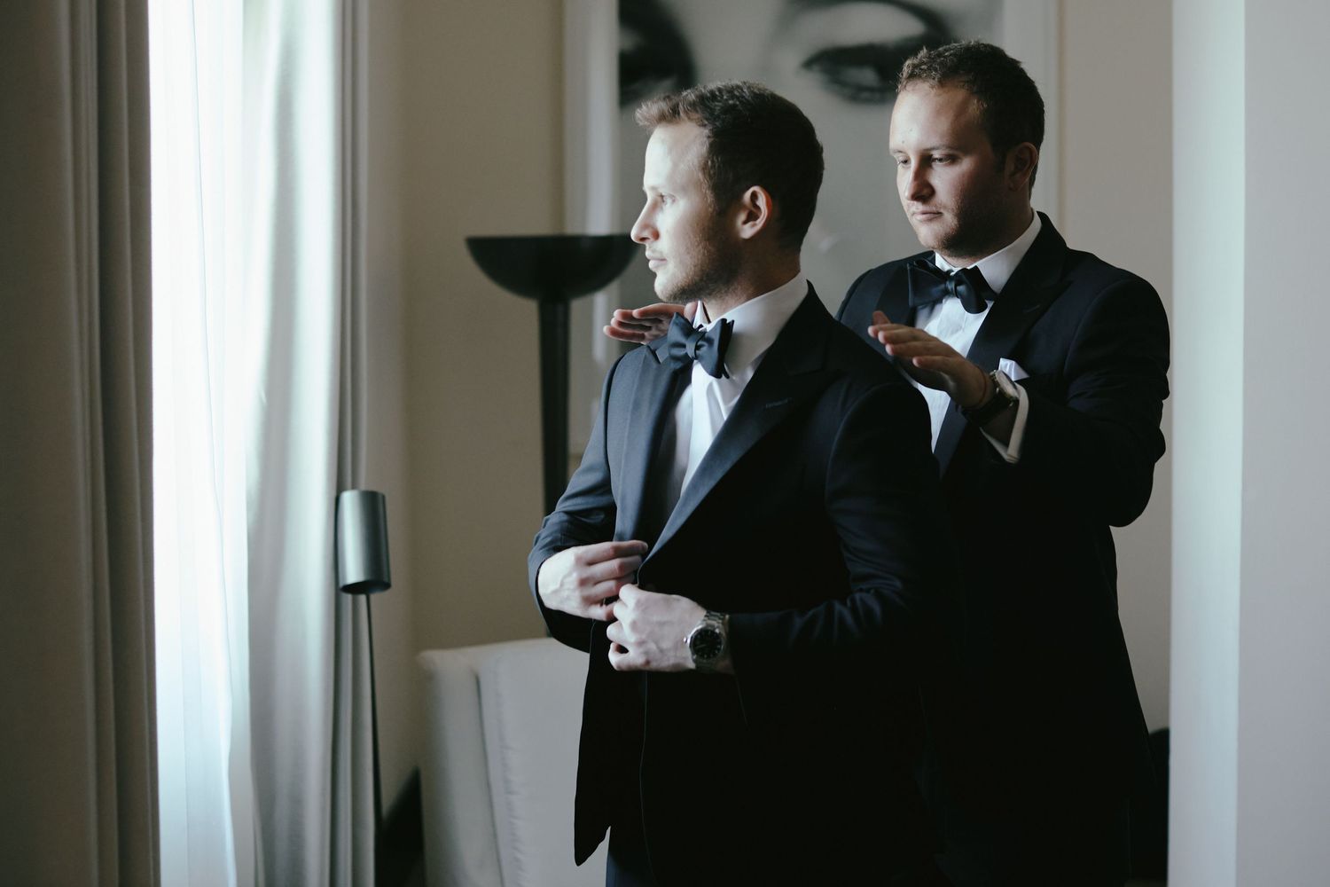 Two people in black tuxedos and bow ties getting ready by a window in a formal setting.