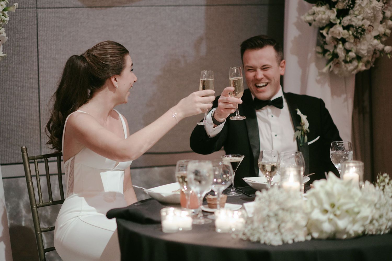 A wedding couple toasts with champagne glasses at a candlelit reception table decorated with white floral arrangements.