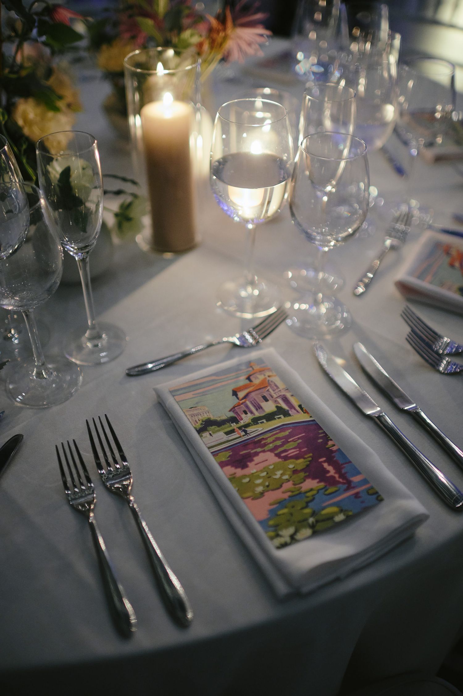 Elegant table setting with wine glasses, cutlery, candle and colorful artistic menu card on white tablecloth.