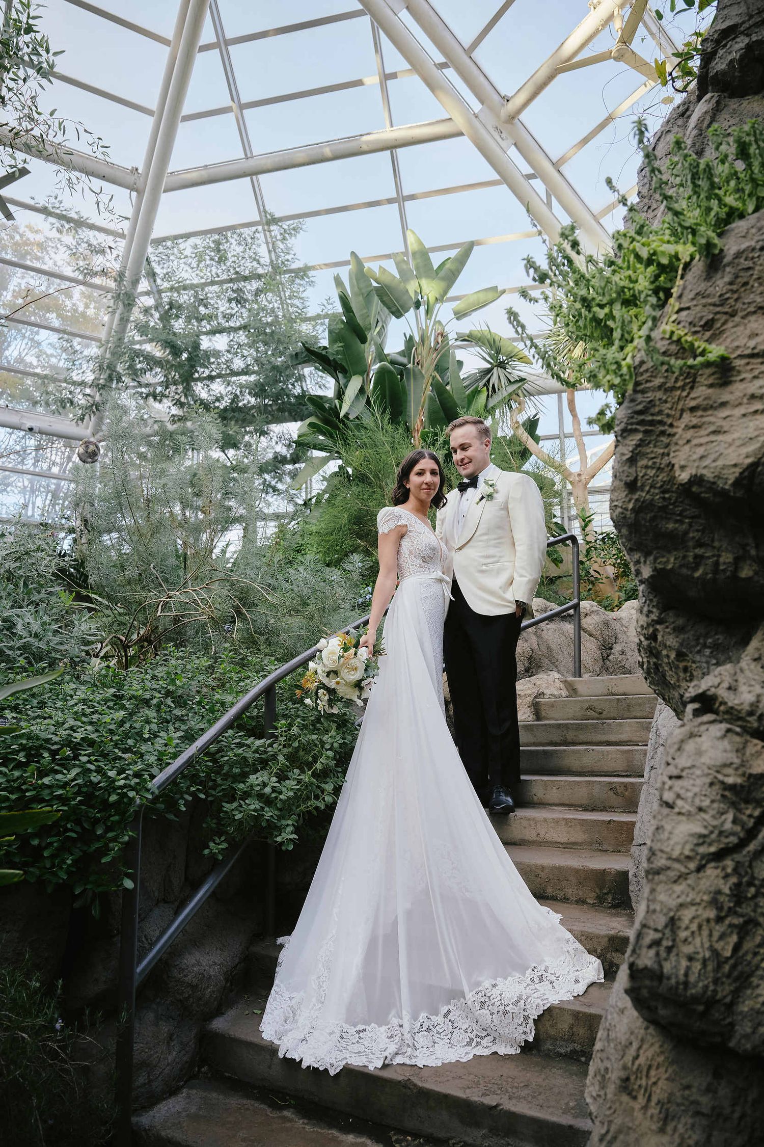 A couple stands on stone steps inside a glass-ceiling conservatory filled with lush greenery and natural light.
