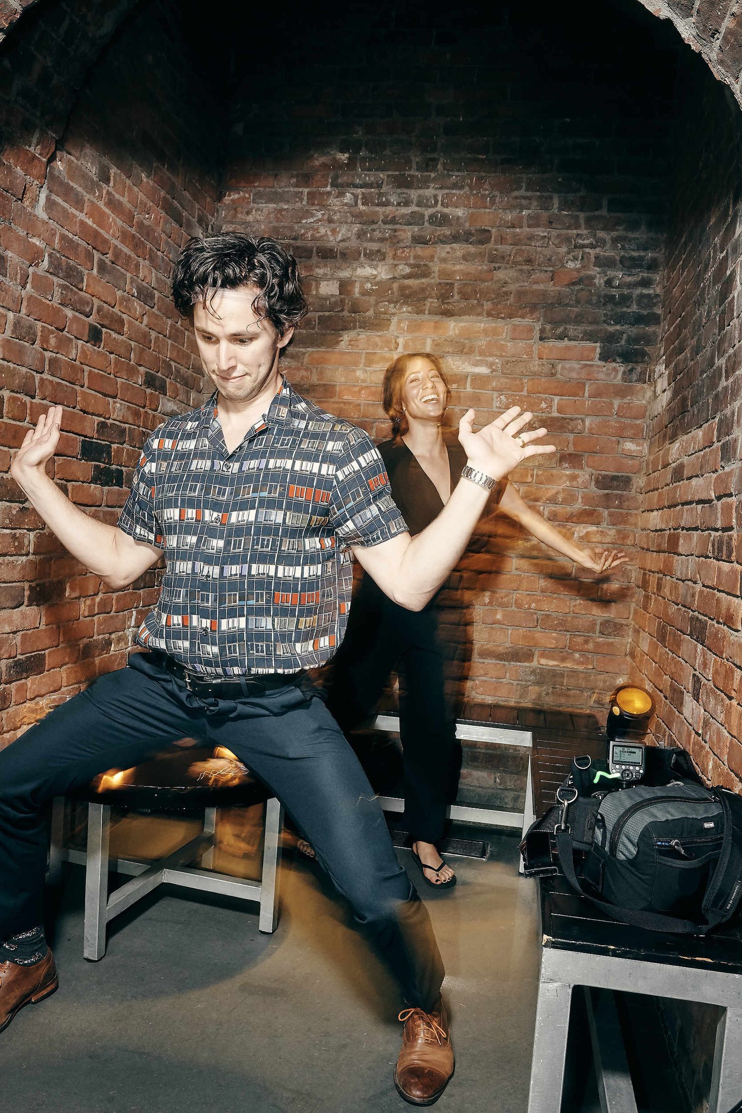 Two dancers strike playful poses against a brick wall in a backstage area with bags and equipment nearby.