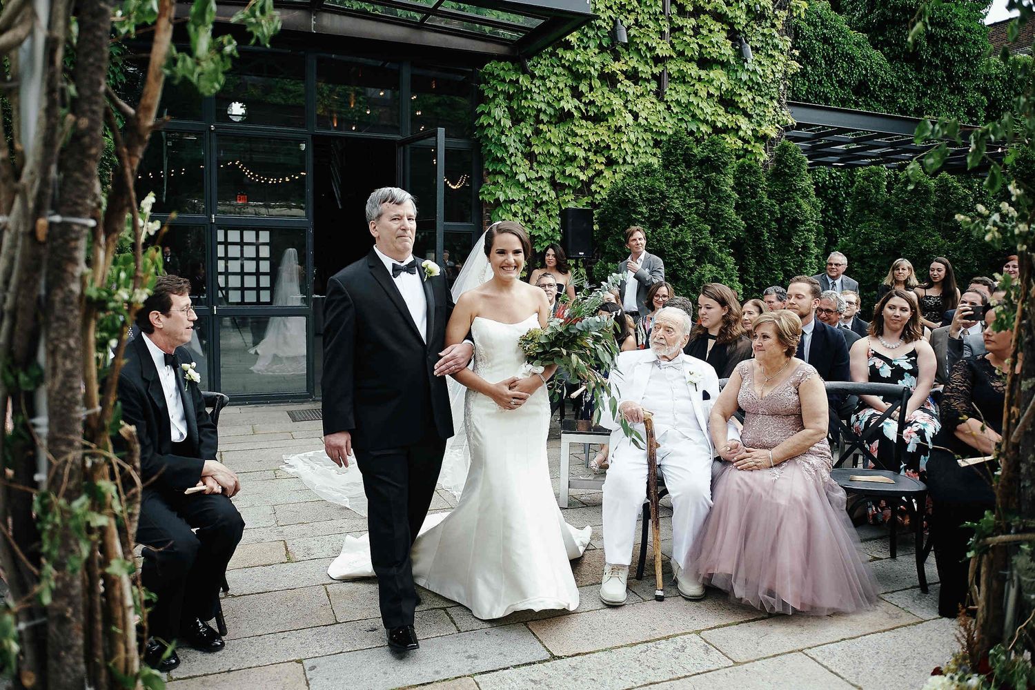 A wedding procession walks along a garden path lined with guests during an outdoor ceremony.
