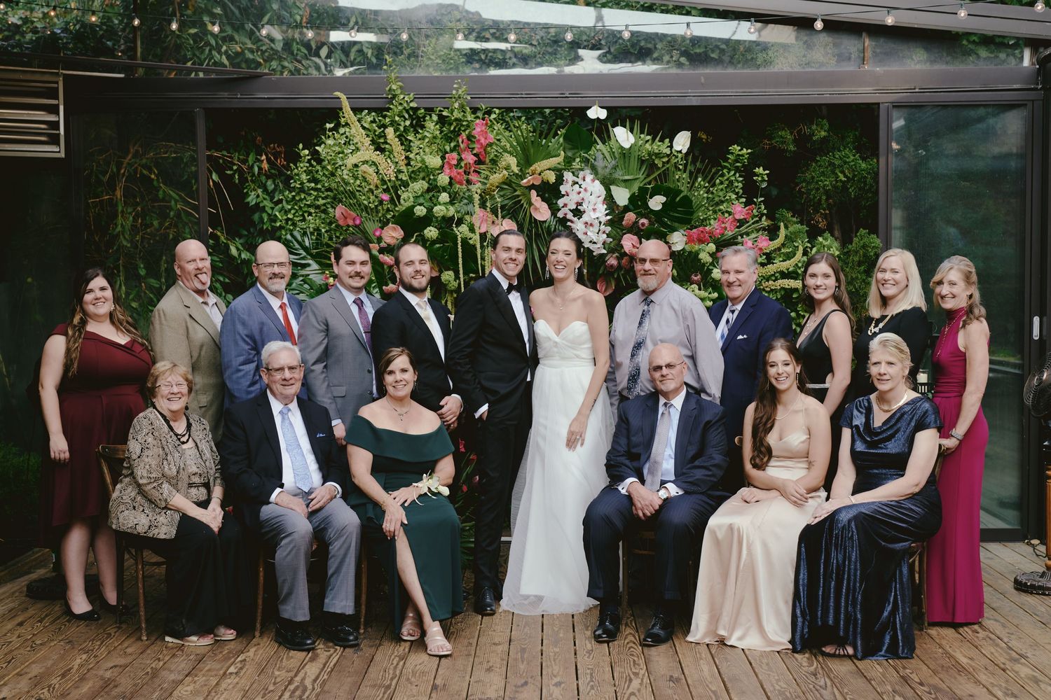Large wedding party stands together for formal group photo on wooden deck with floral decorations.