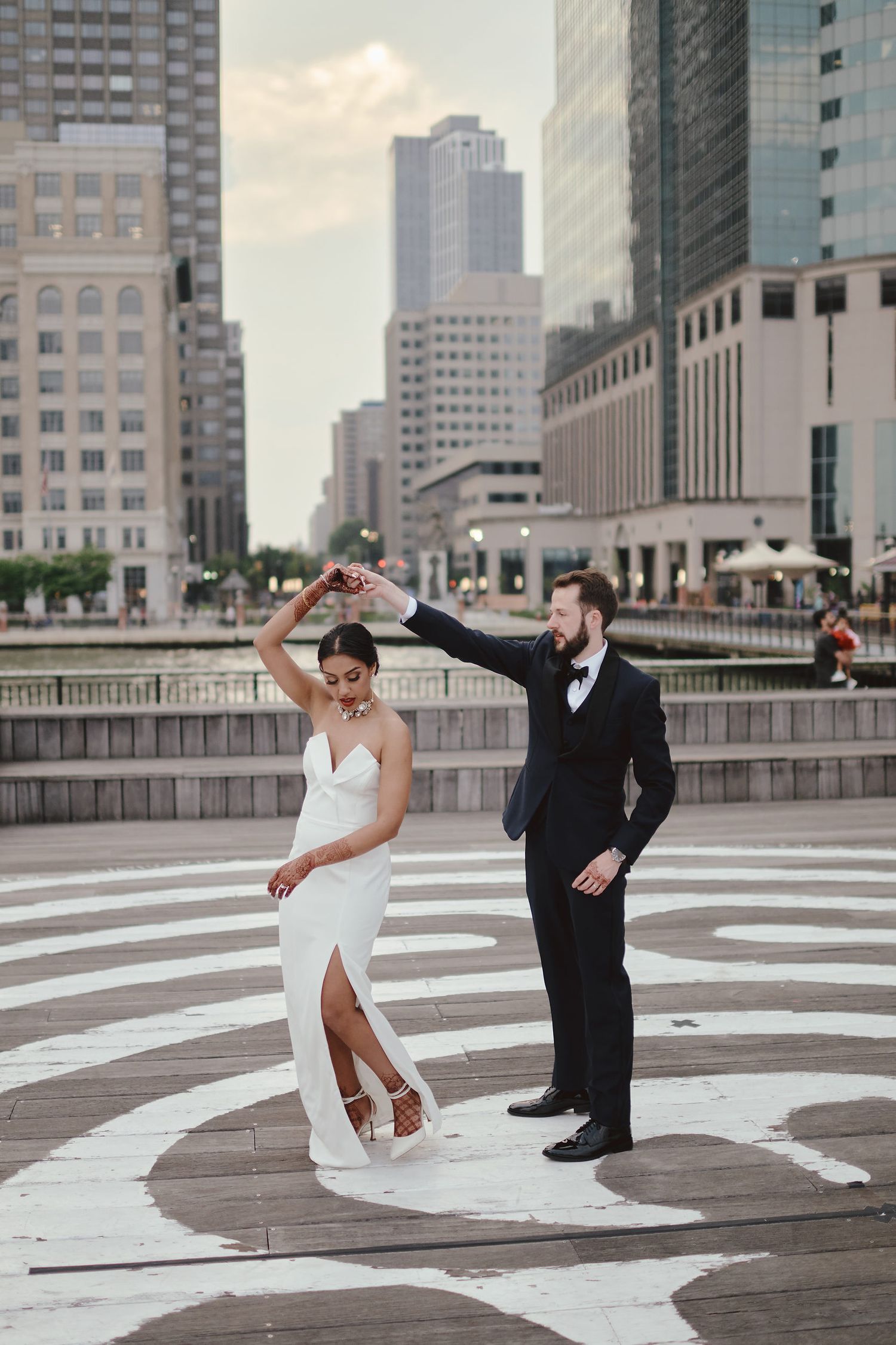 A couple in wedding attire dancing together on a patterned plaza with city skyscrapers in the background.