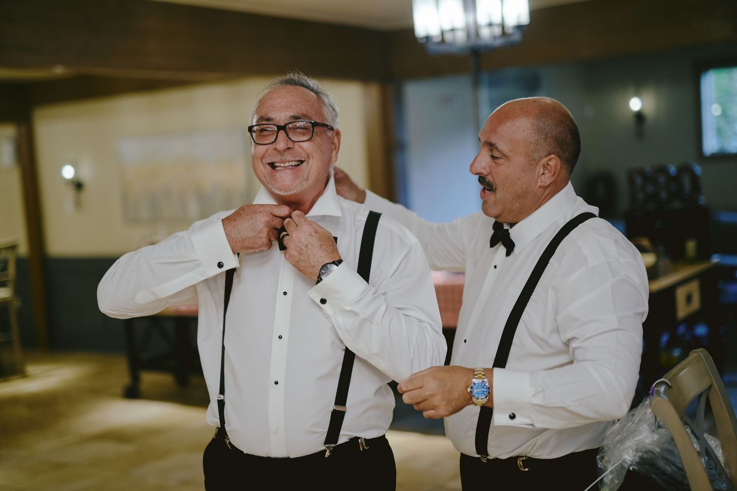 Two men in white dress shirts and suspenders laugh together during a formal event.