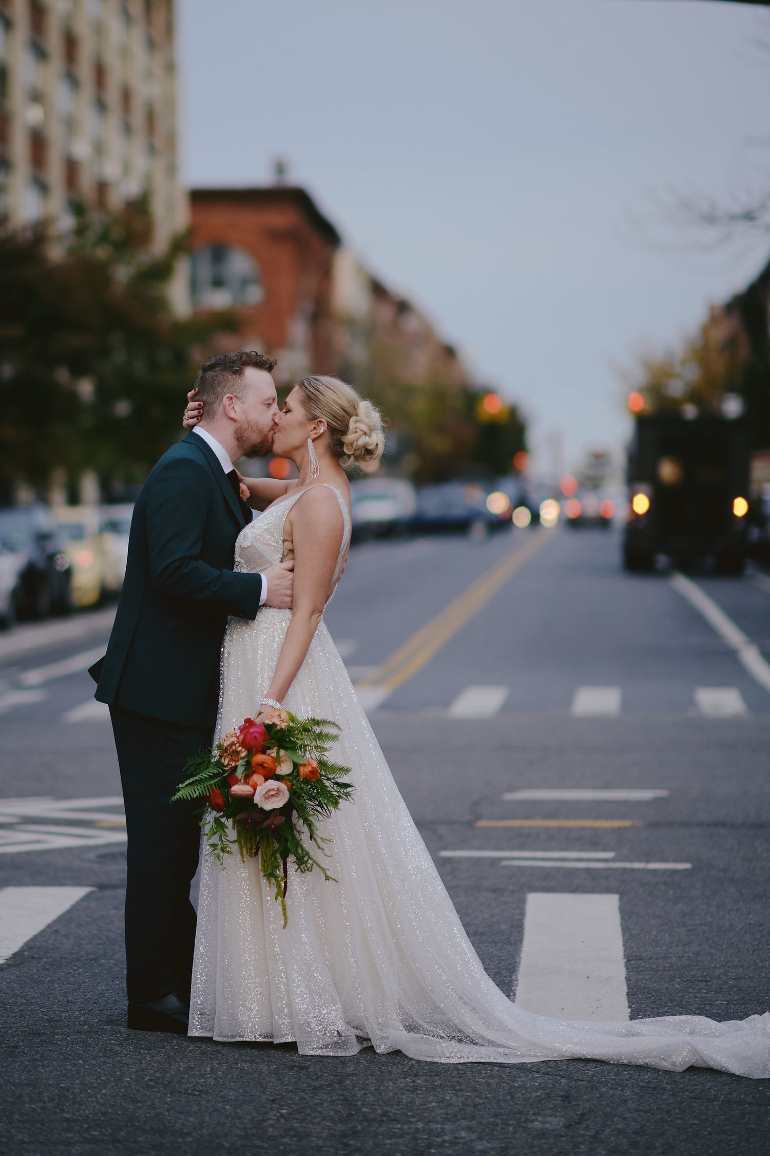A romantic couple shares a kiss on an urban street at dusk while holding a vibrant autumn bouquet.