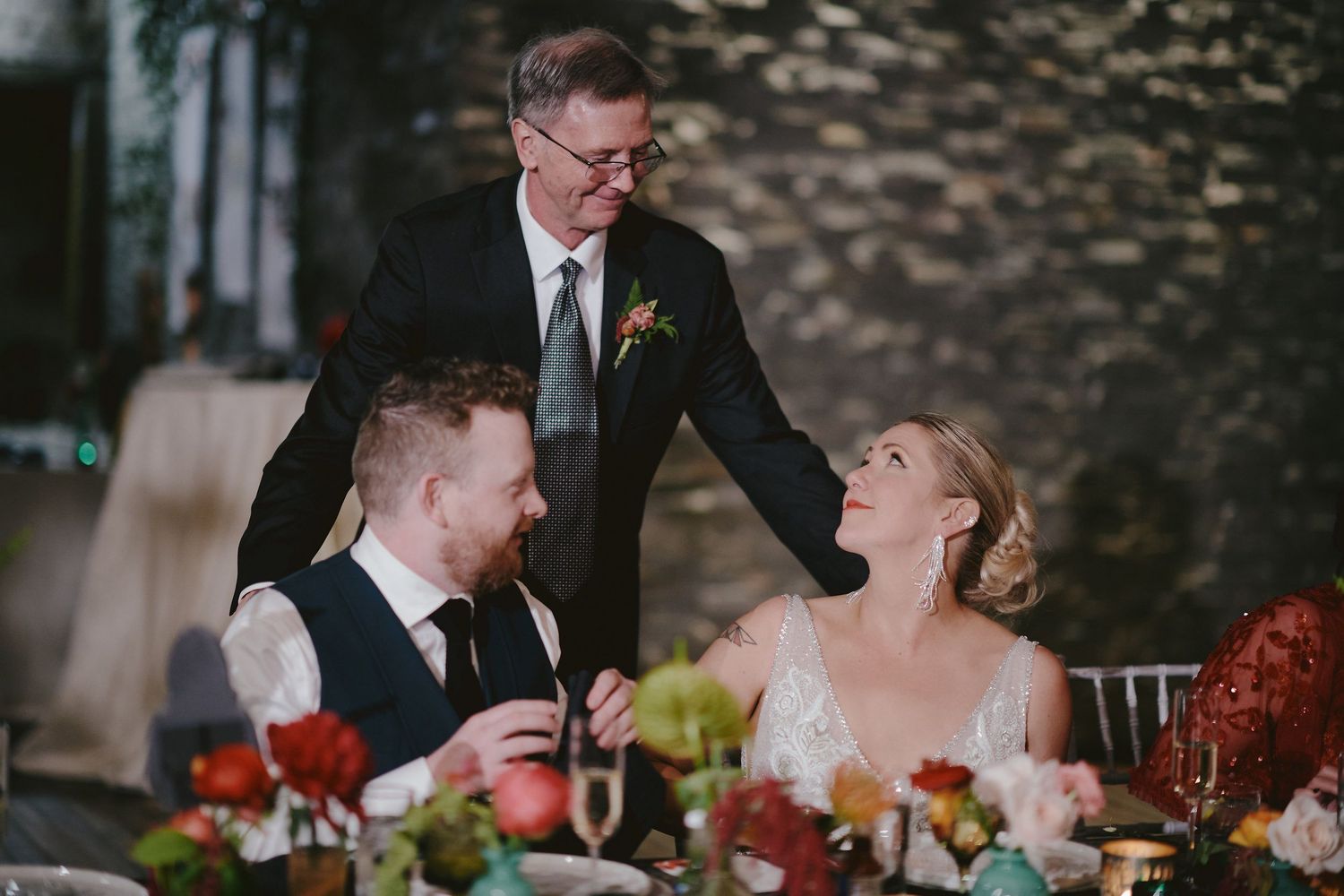 Wedding reception scene with guests sharing joyful laughter at dinner table decorated with red floral centerpieces.