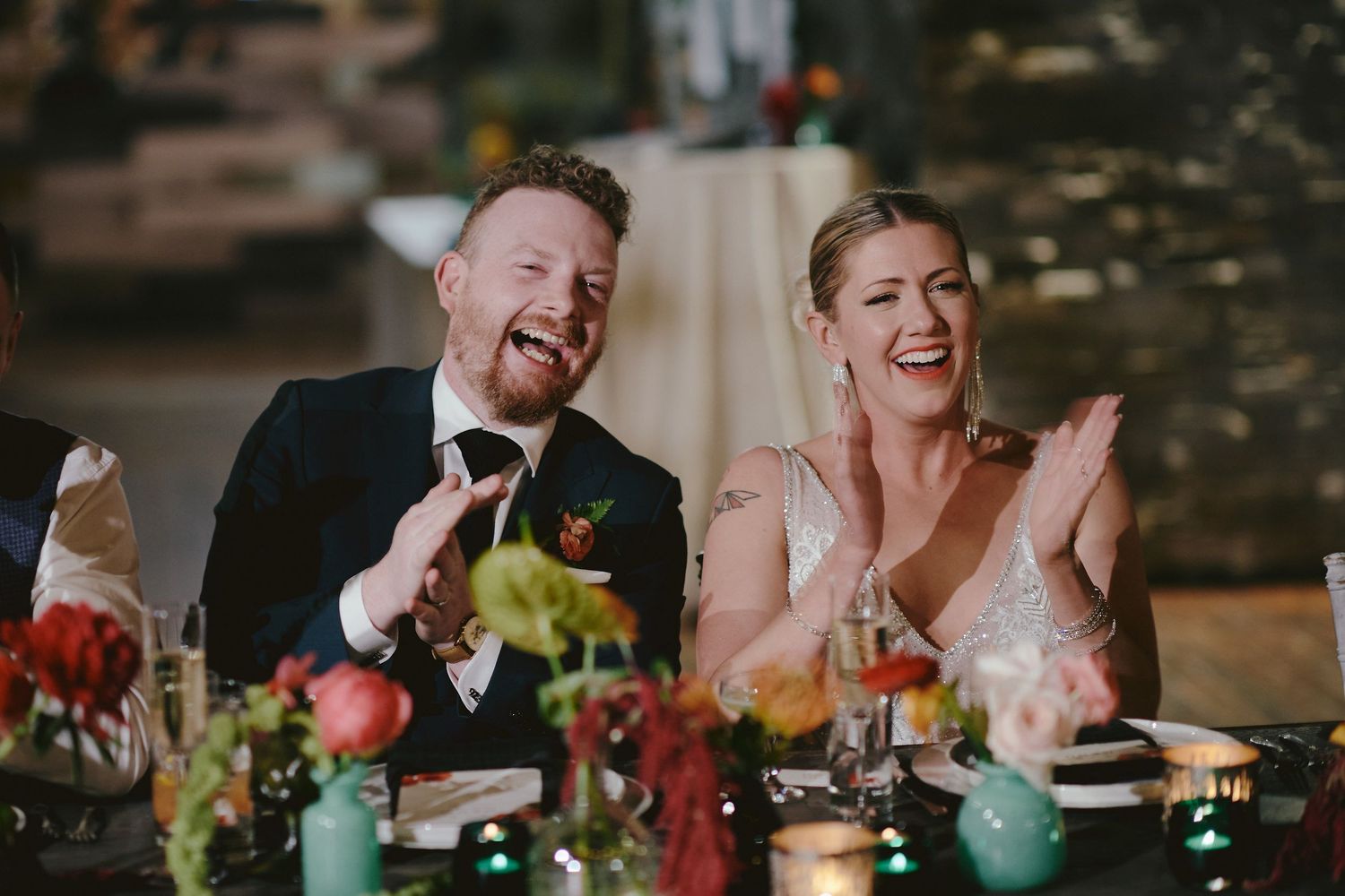 A bride and groom share a joyful laugh together at their wedding reception dinner table.