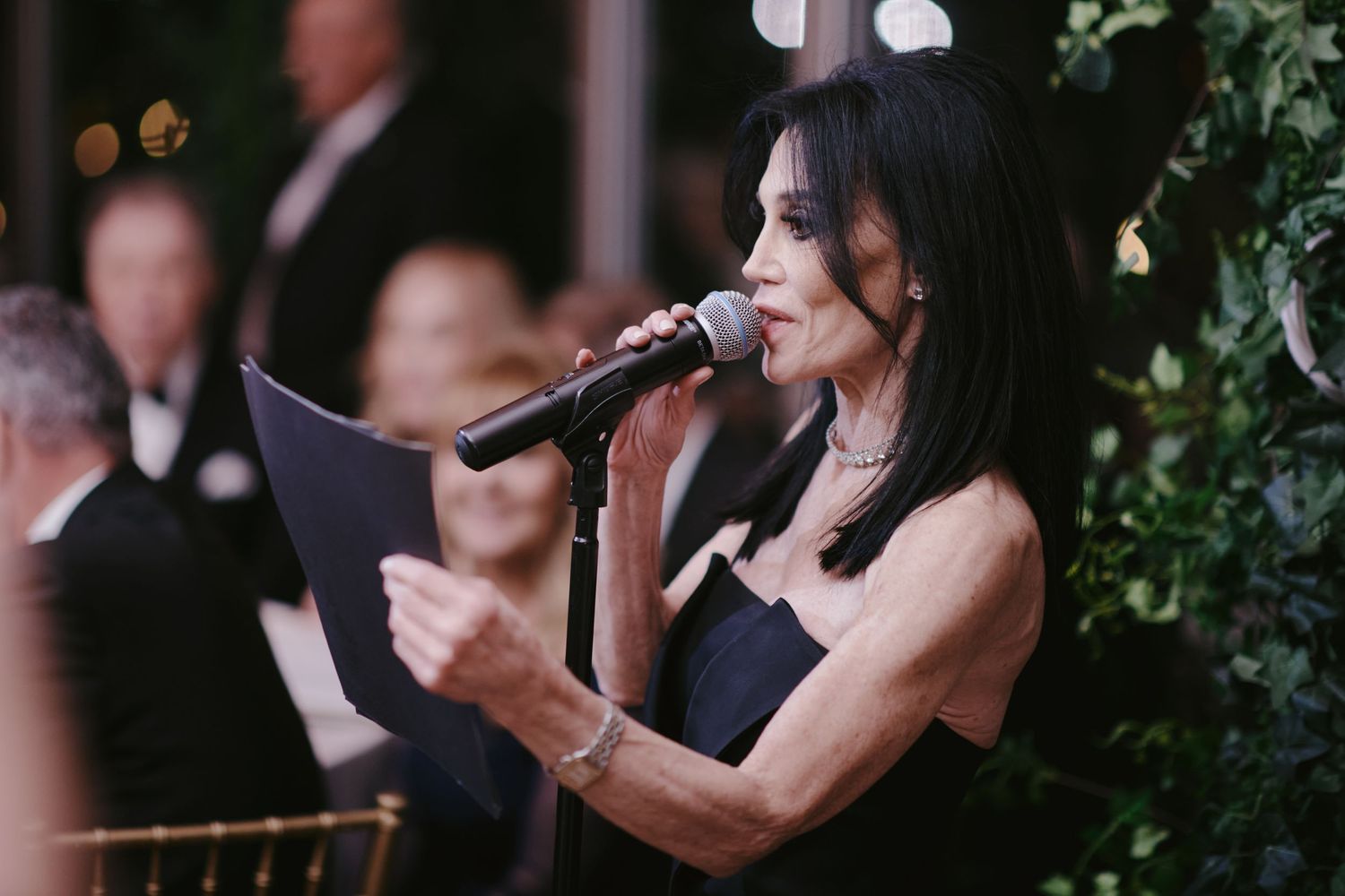 A singer in a black dress holds a microphone and papers while performing at an elegant indoor event.