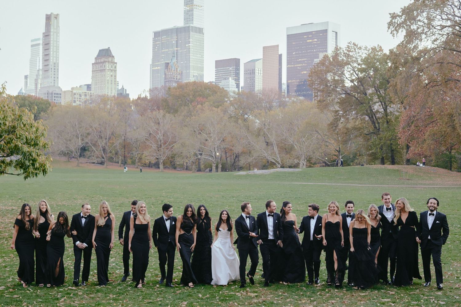 Wedding party group standing in a line on a grassy lawn with New York City skyscrapers rising behind autumn trees.