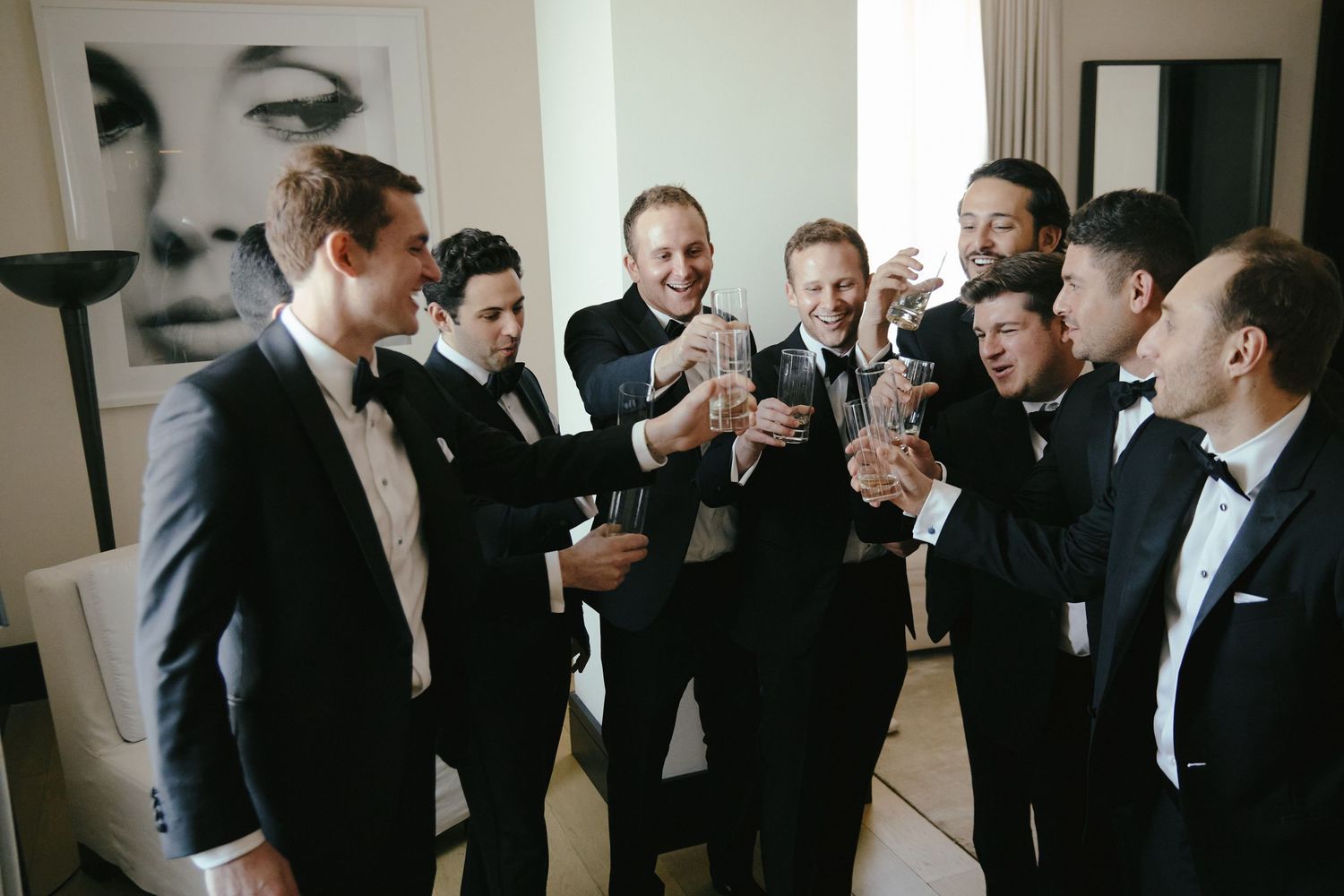 A group of groomsmen in black tuxedos toast with drinks before a wedding celebration.