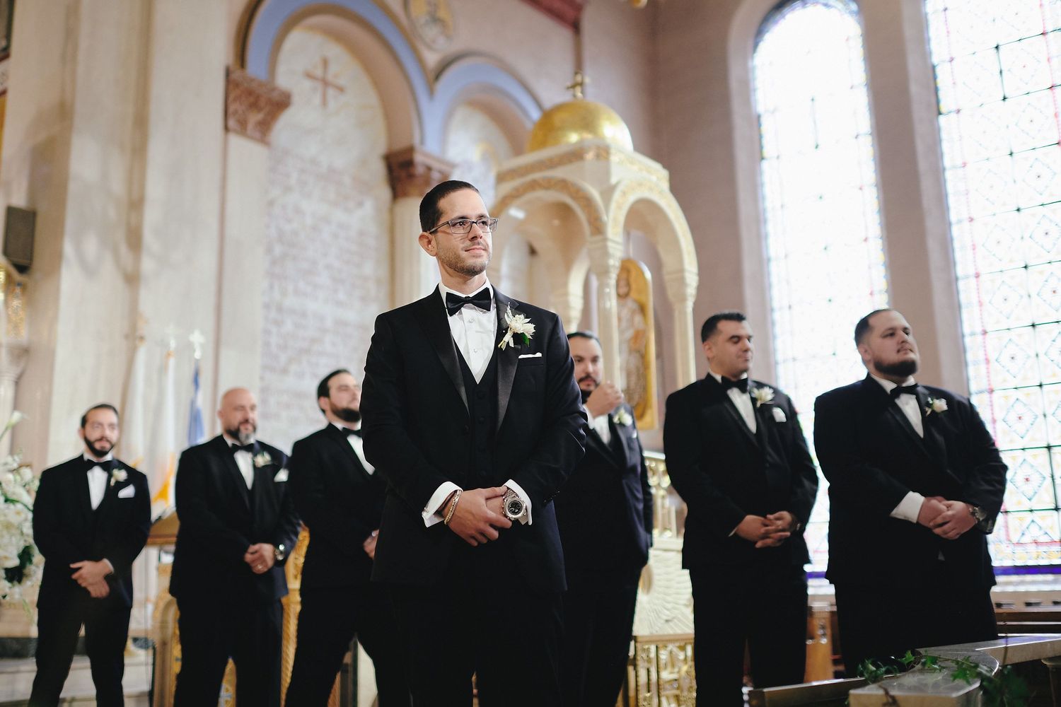 Formally dressed groomsmen stand in a row at the altar inside an elegant church with arched windows and columns.