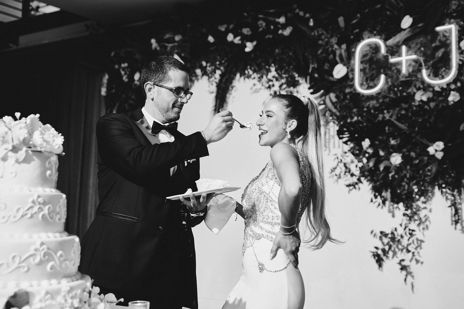 Two newlyweds enjoy a playful cake cutting moment under a decorative floral arch at their wedding reception.