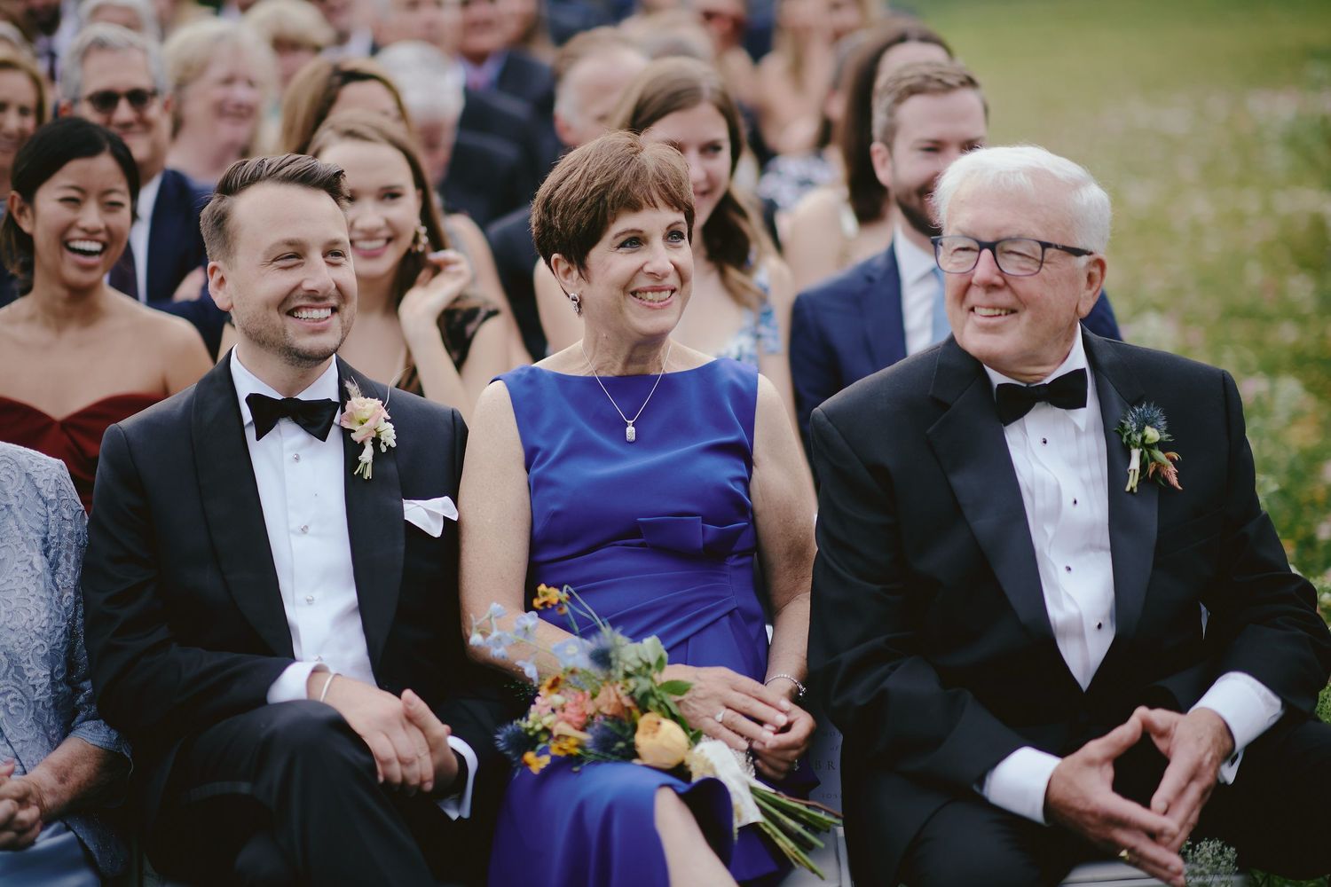 Wedding guests dressed in formal attire smile and laugh while seated outdoors at a ceremony.