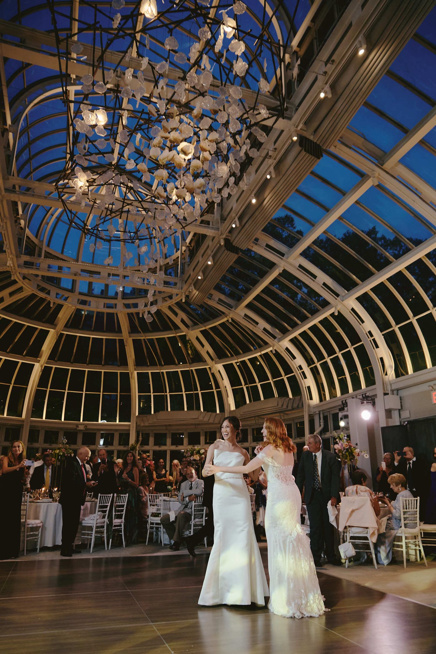 Wedding celebration under a stunning glass dome conservatory with elegant butterfly chandelier and couples dancing on wooden floor.