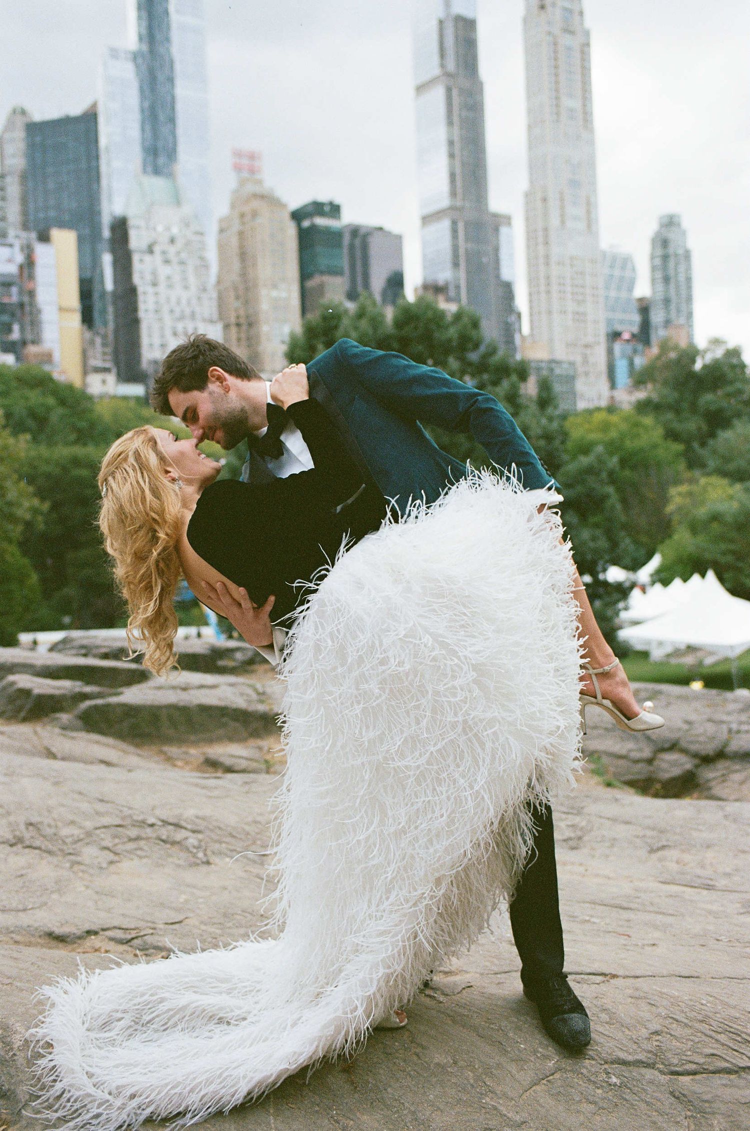 A romantic wedding photo shows a couple kissing dramatically against the New York City skyline in Central Park.