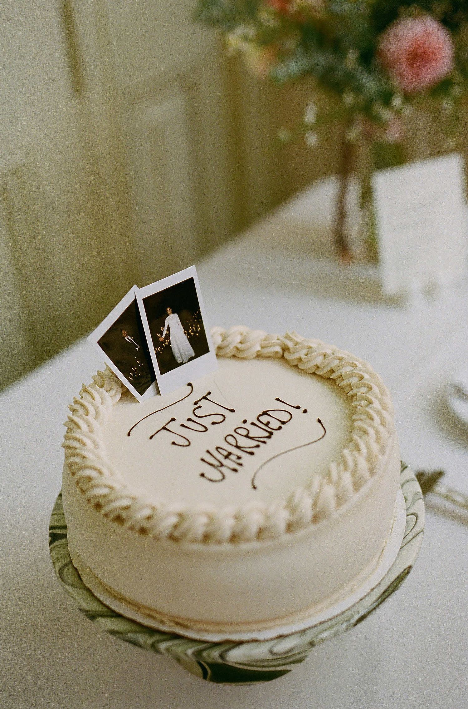 White frosted wedding cake decorated with polaroid photos on top and sitting on a glass cake stand with flowers in background.