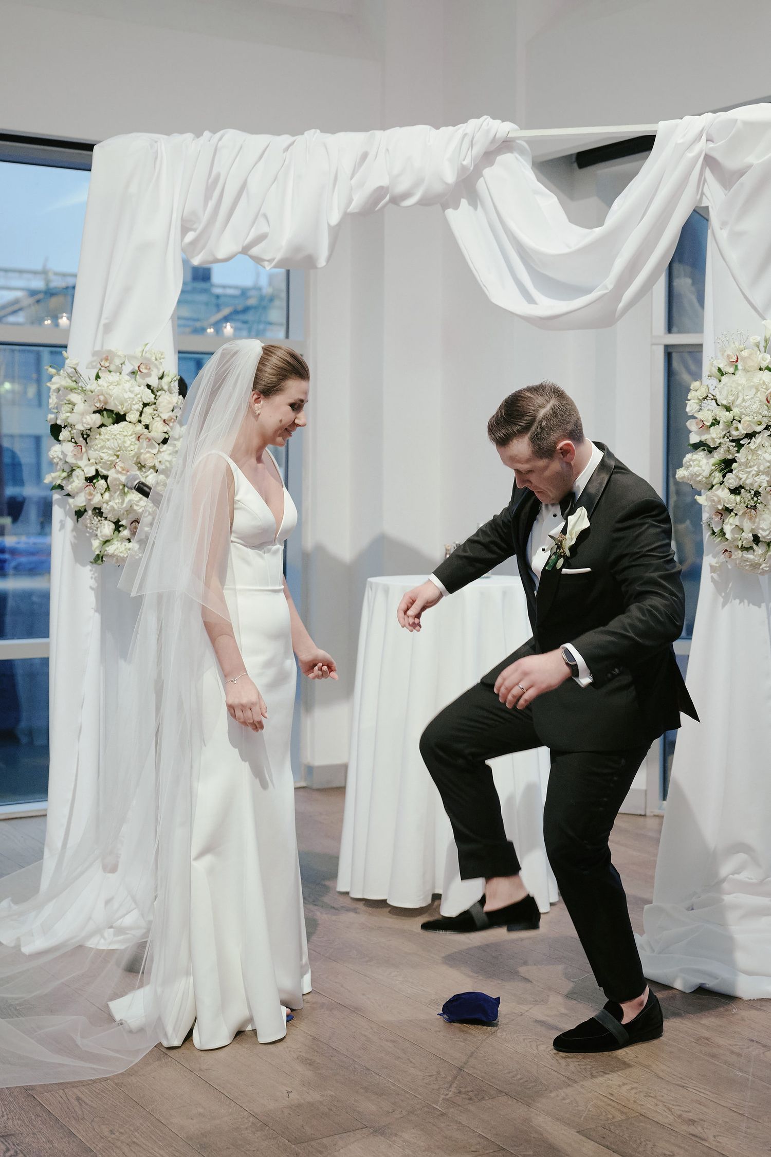 A wedding couple performs the traditional Jewish tradition of breaking the glass under a white draped chuppah with floral arrangements.