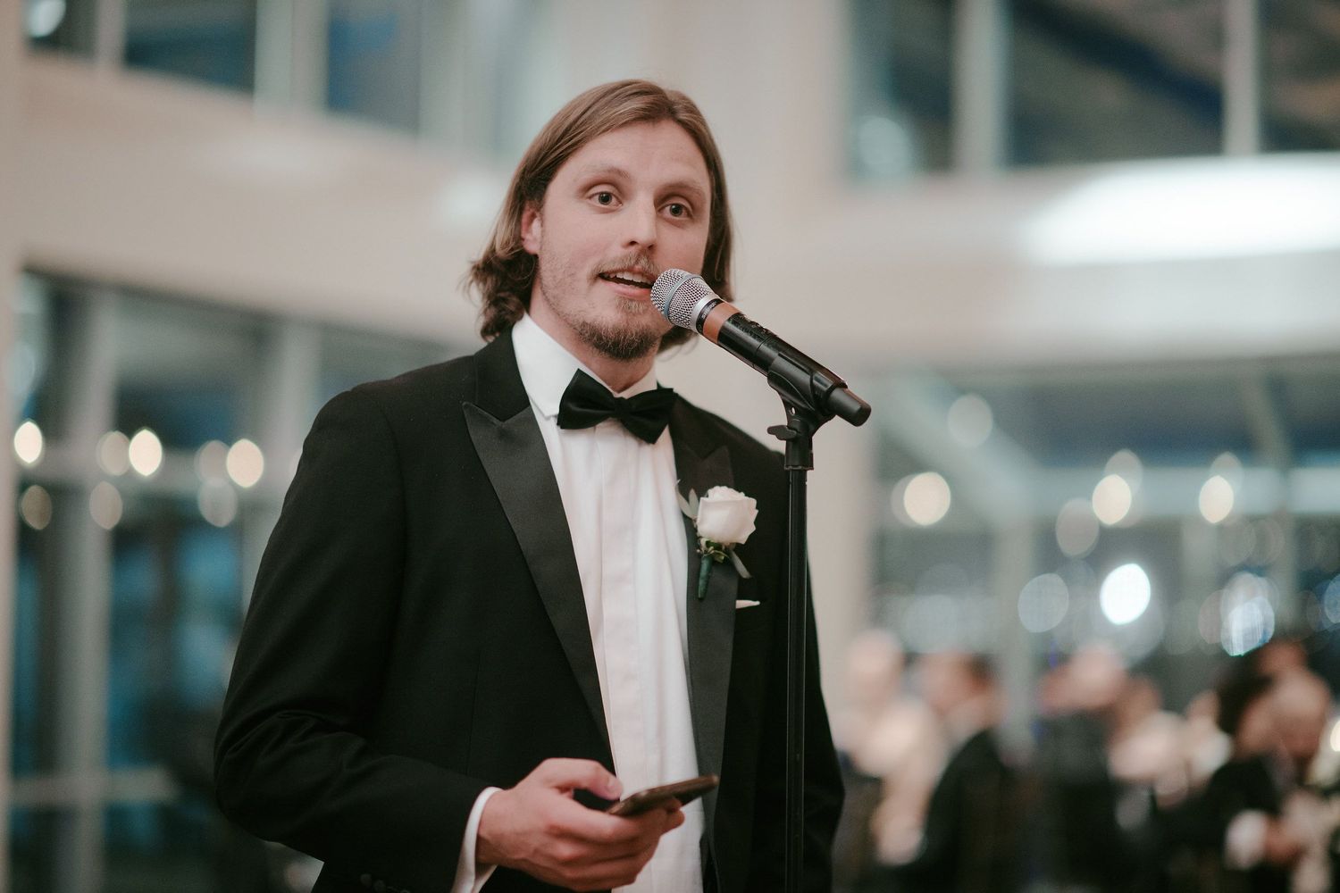 A speaker holds a microphone while giving a speech at a formal event, wearing a black tuxedo with a white boutonniere.