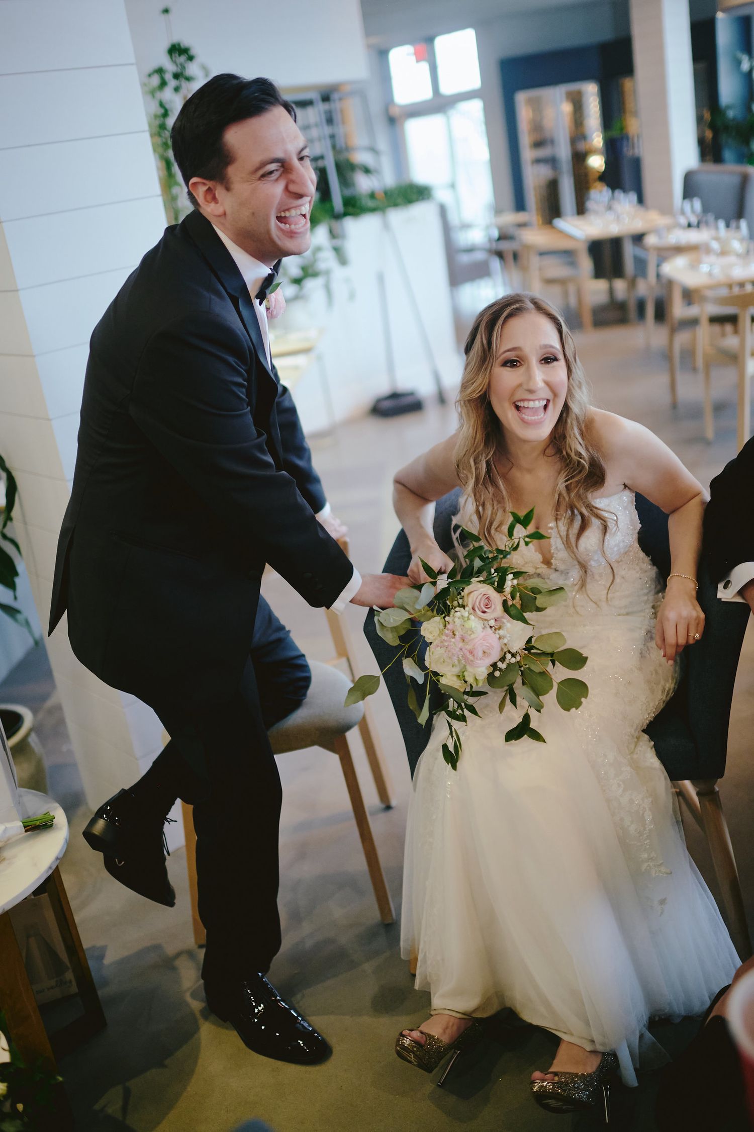 A joyful bride in a white wedding dress and groom in a black suit share a candid moment in a bright reception venue.