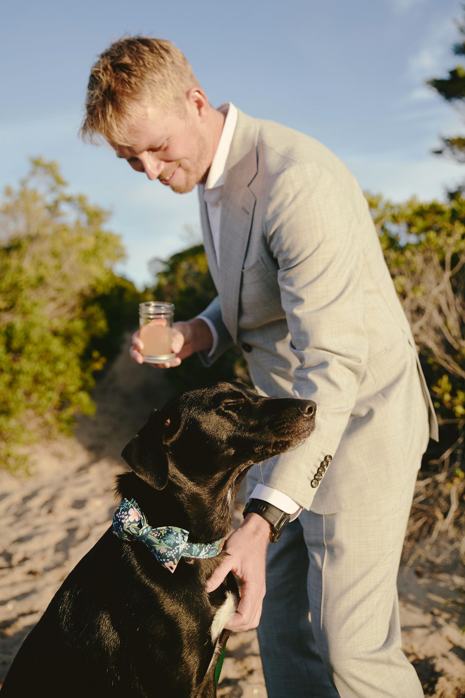 A person in a light gray suit stands outdoors with a black dog wearing a bowtie collar while holding a drink.