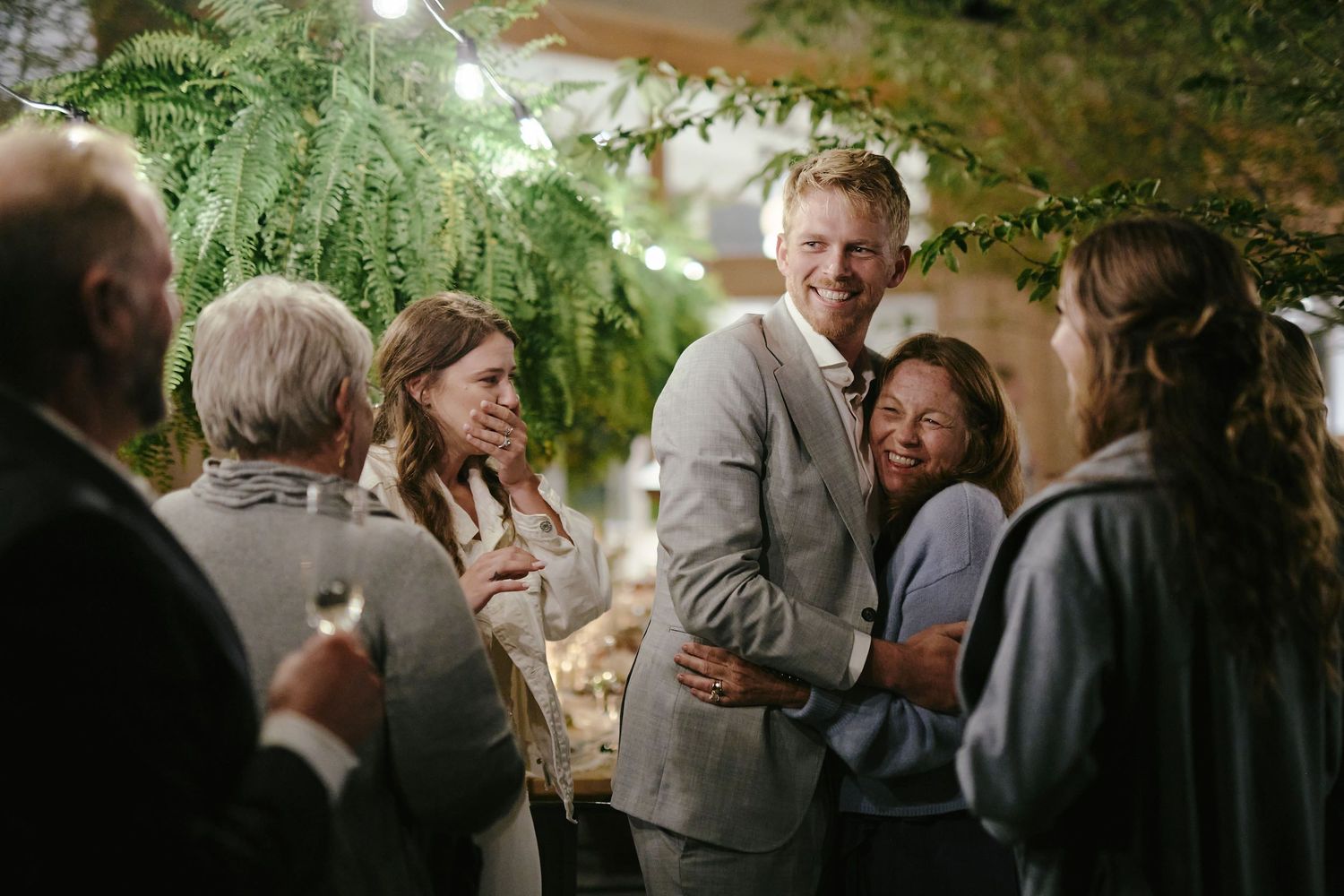 A group of guests mingle and socialize at an evening outdoor gathering under warm lighting and greenery.